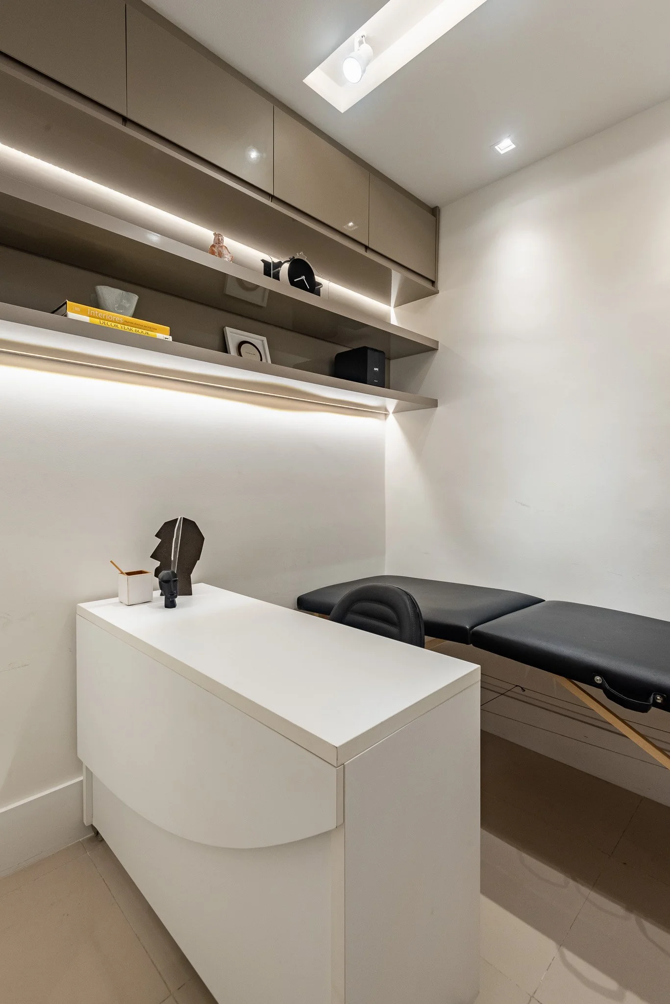 Sala de atendimento com mesa branca, cadeira preta, suporte de exame médico, prateleiras de tom bege, objetos de decoração e iluminação moderna