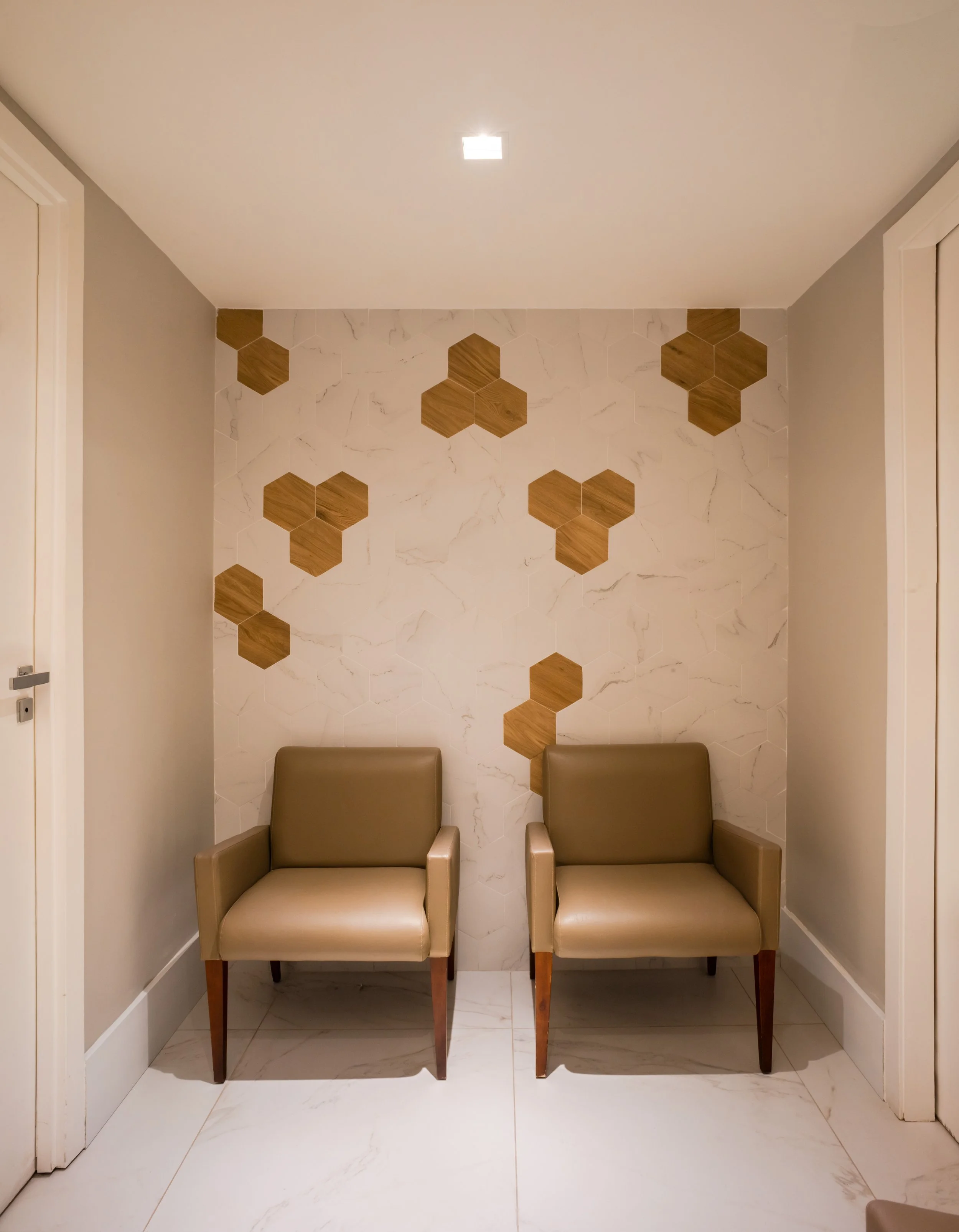 Duas cadeiras de acordo de couro bege em uma parede decorada com mosaico de azulejos brancos e hexagonais de madeira, na parede de fundo há um teto com luz embutida.