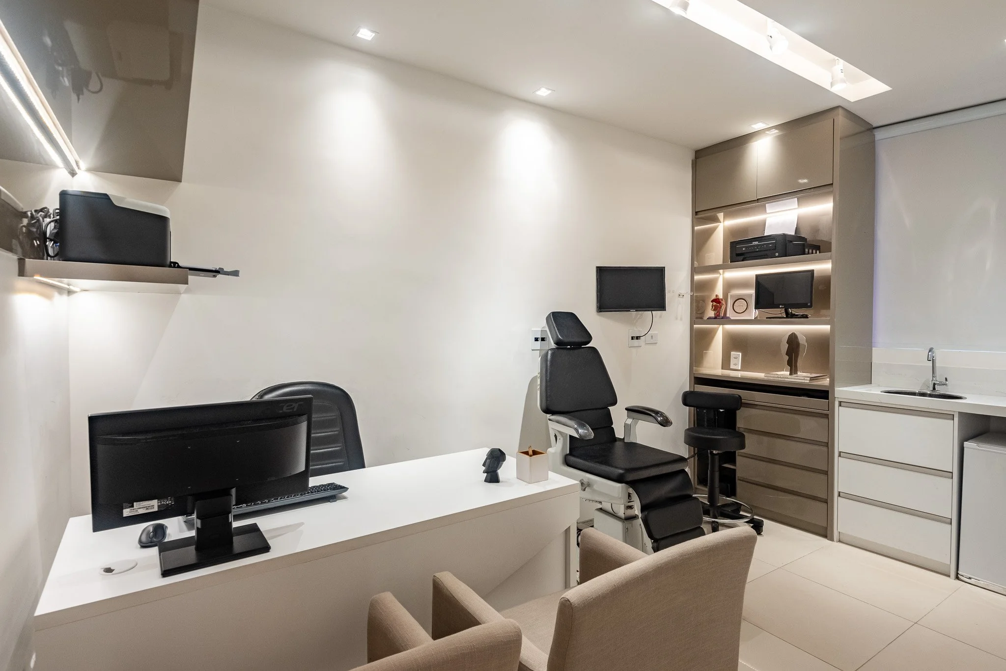 Consultório médico com cadeira de exame, computador, armários, televisão, prateleiras com objetos decorativos e uma pia.