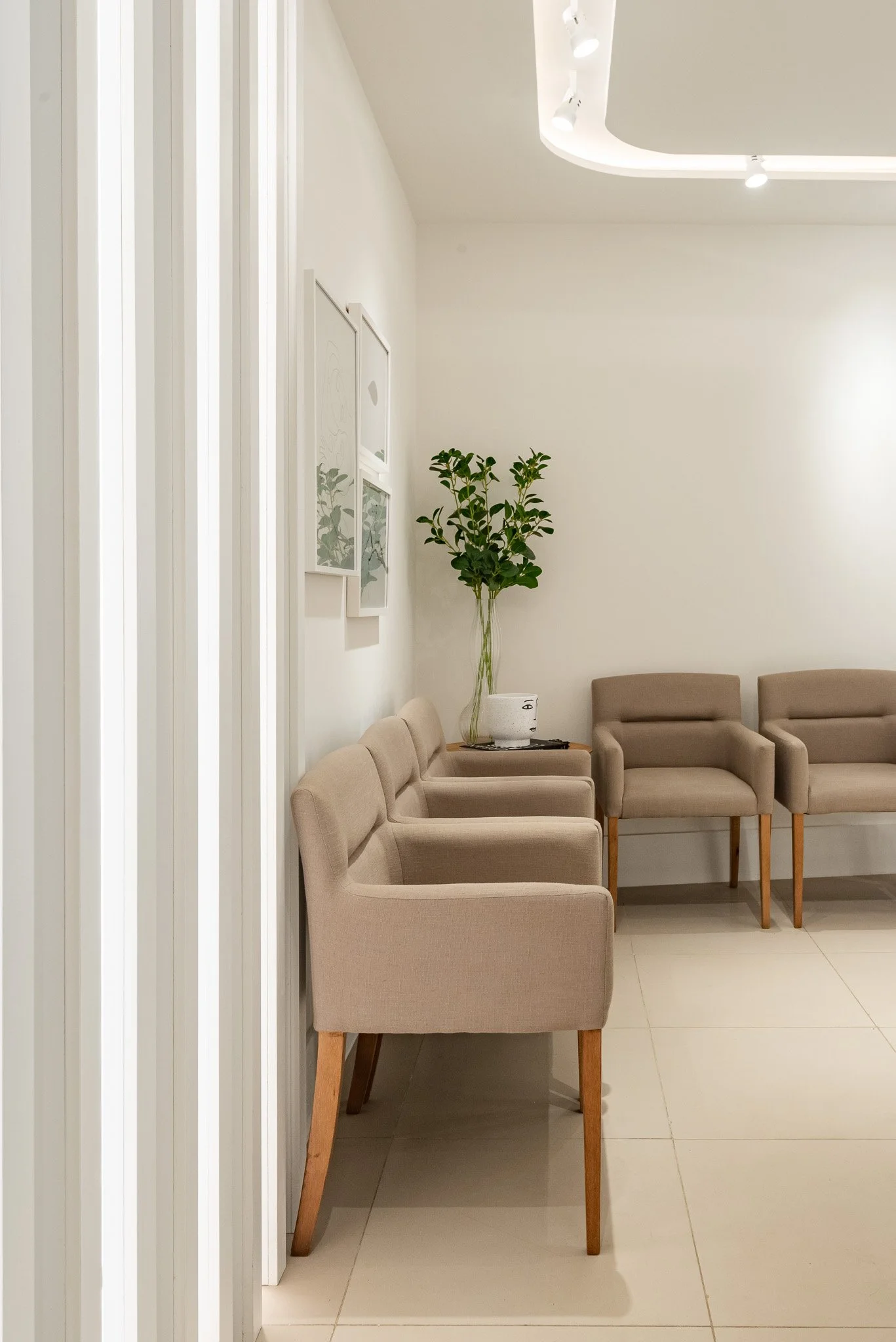 Sala de espera com cadeiras bege, uma planta grande e quadros na parede, iluminação moderna no teto.