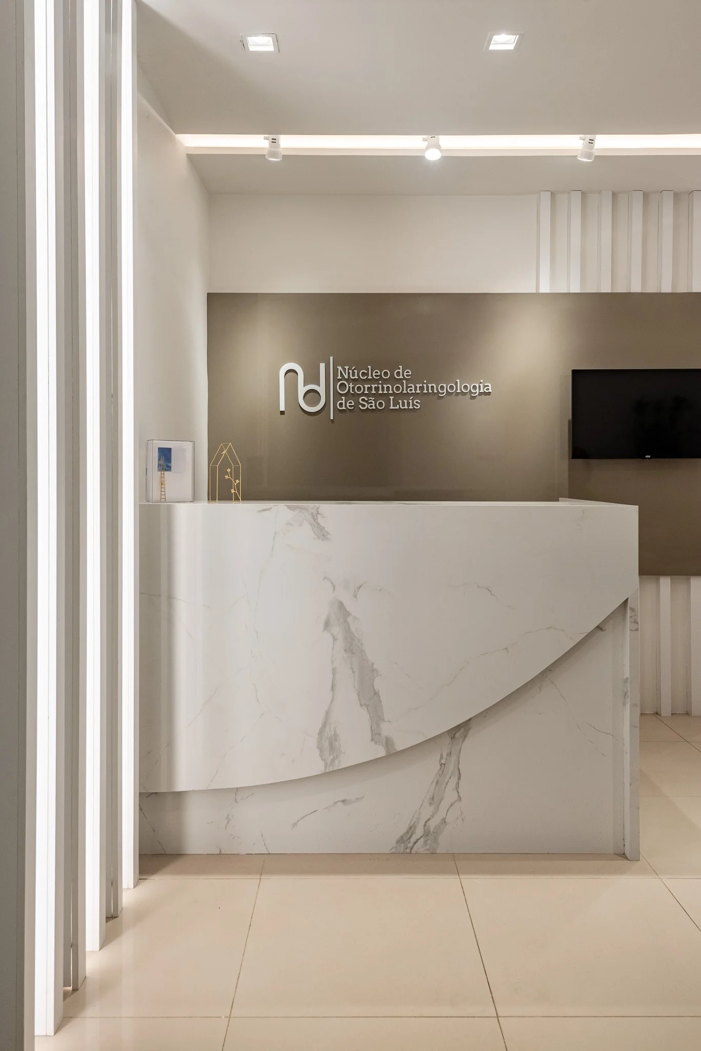 Recepção de clínica médica com parede cinza e painel branco de mármore na bancada. Logo da clínica 'Núcleo de Otorrinolaringologia de São Luís' na parede ao fundo ao lado de um monitor de TV desligado.