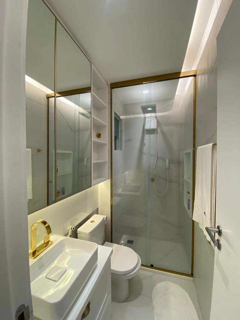 Banheiro moderno com pia branca, torneira dourada, espelhos amplos, vaso sanitário branco, aquecedor de toalhas, box de vidro com detalhes dourados, paredes de mármore, e uma janela na área do chuveiro.