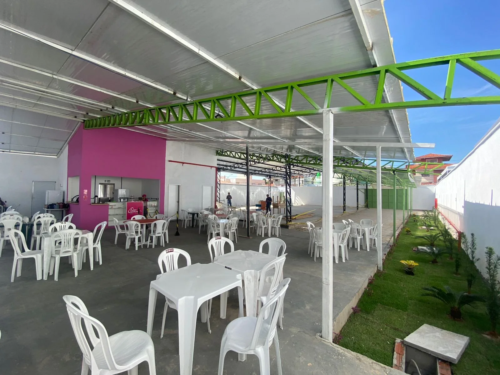 Área de churrasqueira com mesas e cadeiras de plástico branca, espaço coberto com telhado metálico, parede rosa ao fundo, jardim ao lado com plantas e grama, espaço aberto para eventos.