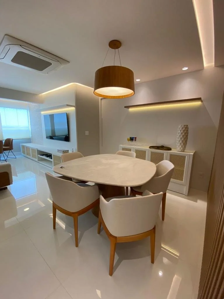 Sala de jantar com mesa de mármore, seis cadeiras brancas de madeira, luminária de teto em formato de cilindro de madeira, decoração minimalista com vaso branco alto e parede com iluminação indireta.