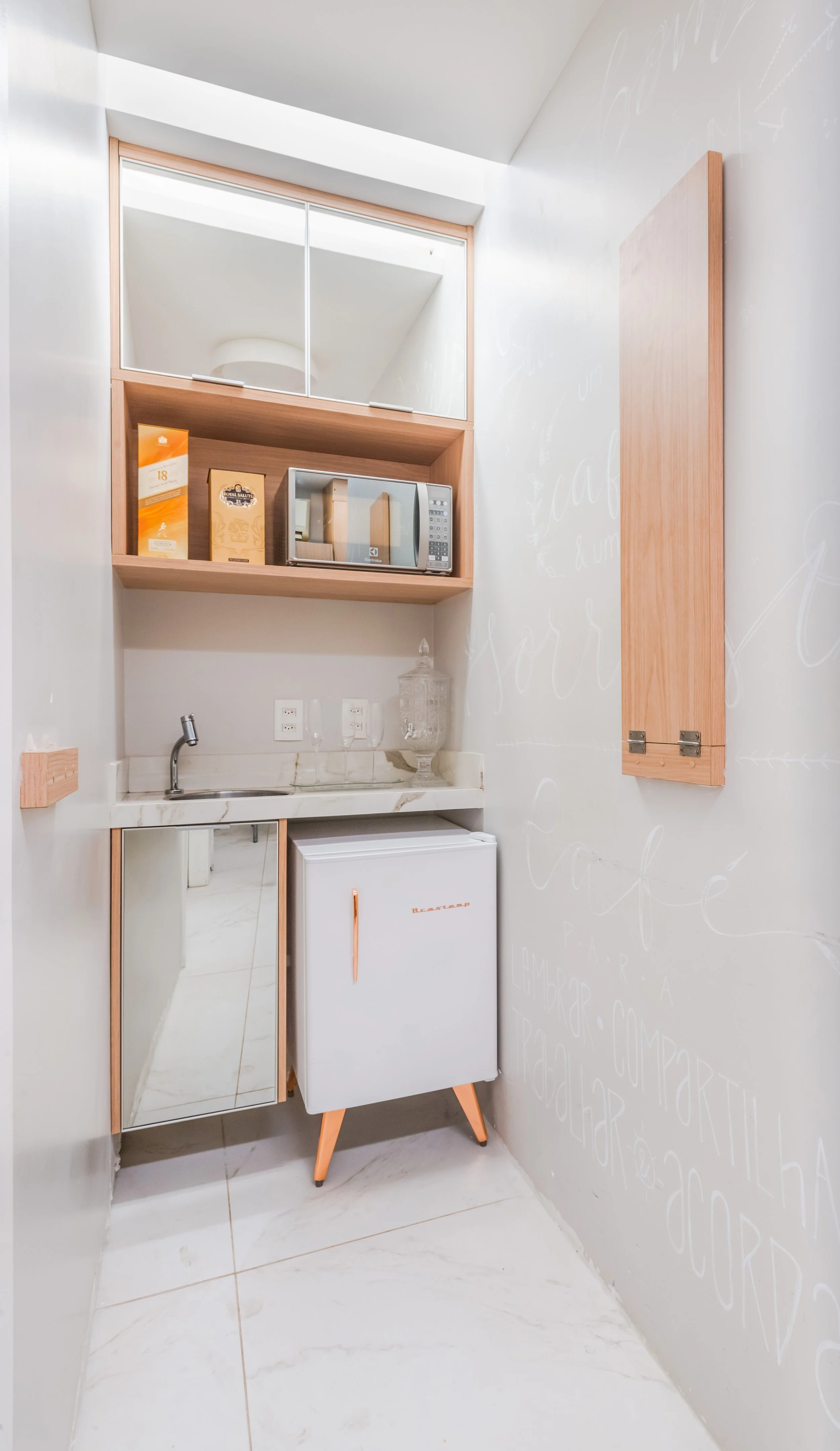 Pequena cozinha ou lavabo com pia, micro-ondas, mini geladeira, armário de madeira, espelho e utensílios diversos.