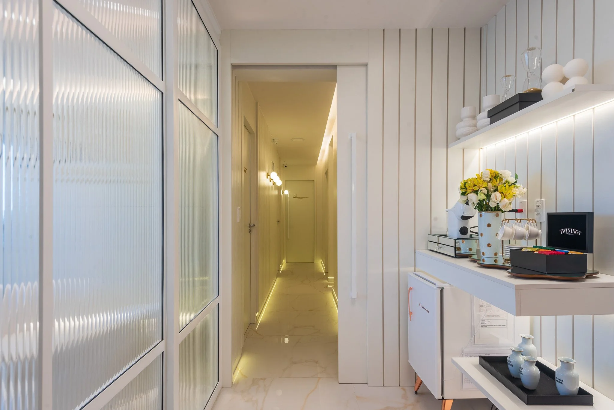 Corredor com paredes de madeira branca, piso de mármore branco, iluminação indireta, decoração com flores e objetos de decoração na parede e na mesa