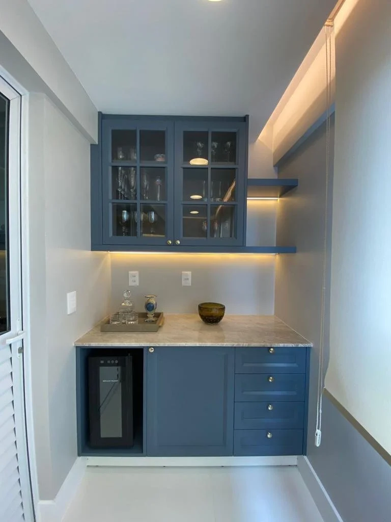 Mini bar com armário azul com portas de vidro, prateleiras internas com taças e copos, e uma bancada de mármore claro, com eletrodoméstico de vinho na parte inferior esquerda, iluminação embutida sob o armário, e uma porta de correr branca à esquerda