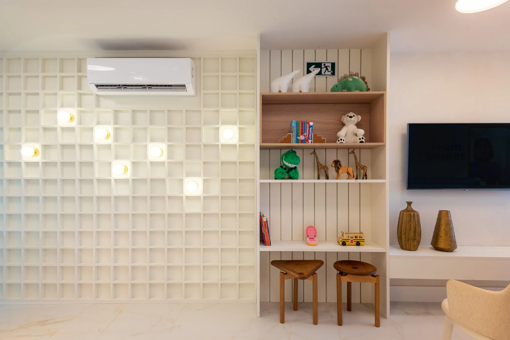 Sala com estante contendo brinquedos, livros e objetos decorativos, parede com painel branco de quadrados e ar condicionado na parte superior