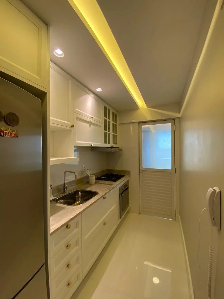 Cozinha estreita com armários brancos, geladeira à esquerda, pia, fogão e forno na bancada, porta de vidro na extremidade, iluminação embutida no teto.