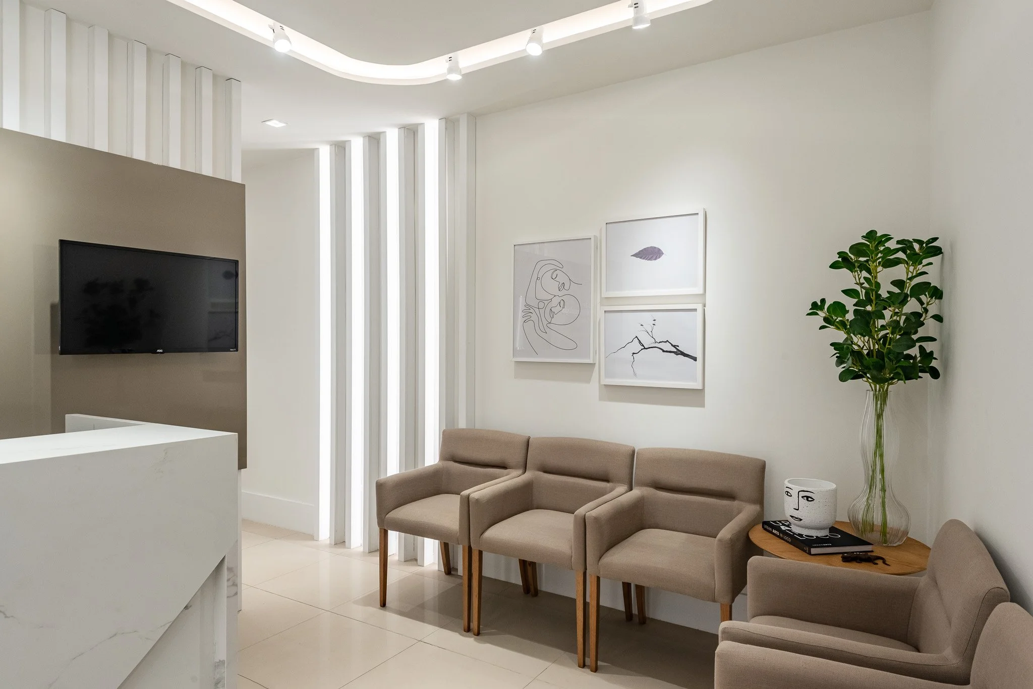 Sala de espera com sofá bege, parede branca com quadros decorativos, planta em vaso na mesa ao lado, televisão e iluminação moderna no teto