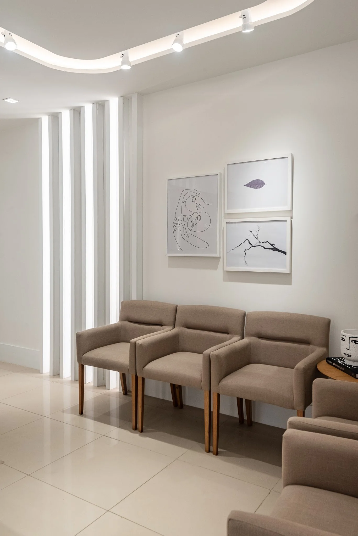 Sala de espera com três cadeiras de tecido bege e pernas de madeira, parede branca com três quadros de arte minimalista, iluminação suave no teto e piso de azulejo claro.