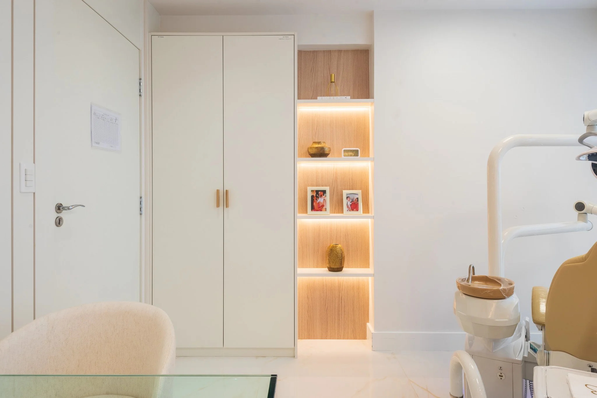 Sala de espera ou consultório, com armário branco, prateleira de madeira com iluminação, vasos decorativos e duas fotos em quadros. Também há equipamento odontológico na direita.
