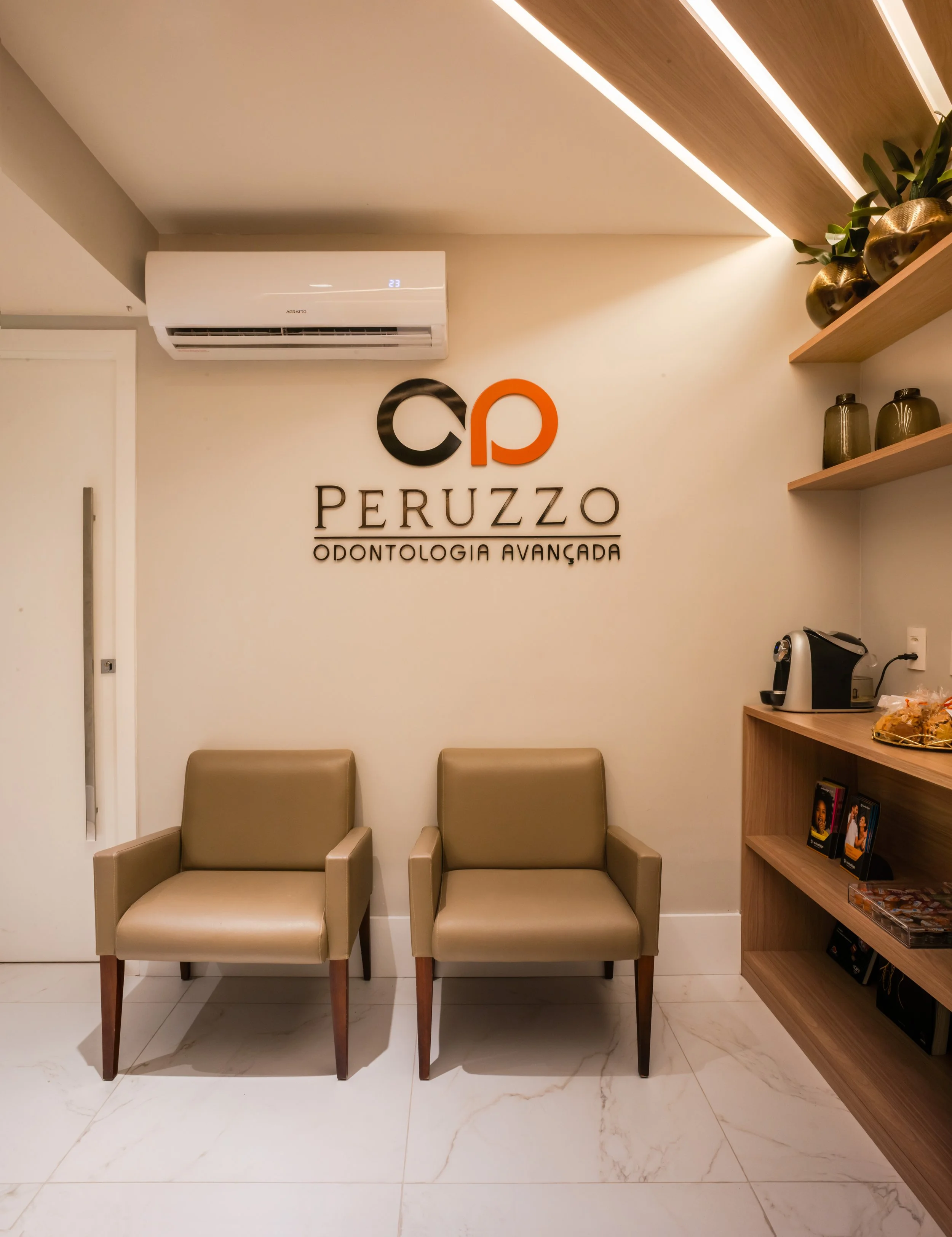 Sala de espera com duas poltronas bege, logo da clínica odontológica Peruzzo e prateleiras de parede com vasos decorativos e livros