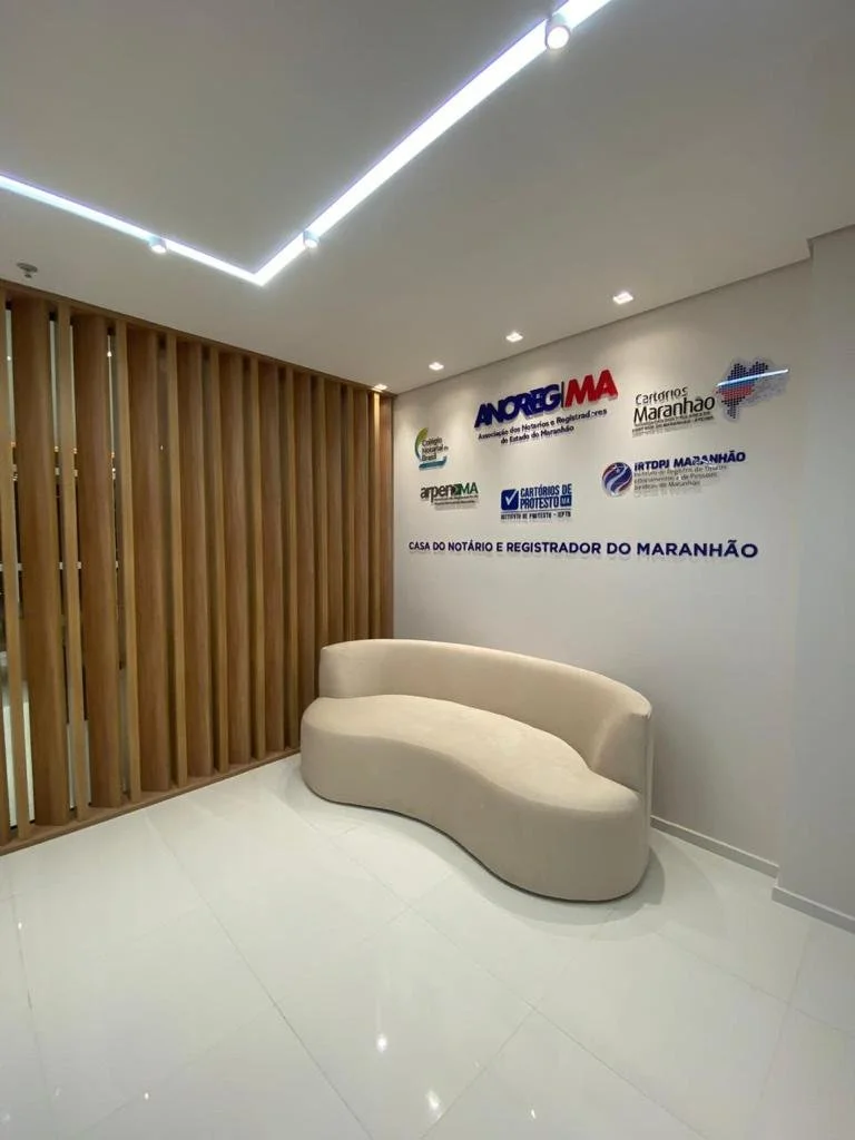 Sala de espera com uma parede de riconhecimento de nomes e certificados do Maranhão, uma bancada de madeira com uma parede branca ao fundo, um sofá curvo bege e iluminação moderna no teto.
