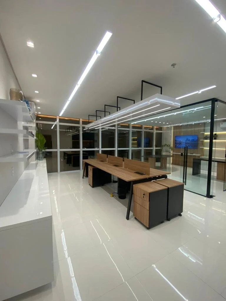 Escritório moderno com mesas de trabalho, prateleiras brancas na parede, piso branco, iluminação LED no teto e paredes de vidro que separam ambientes