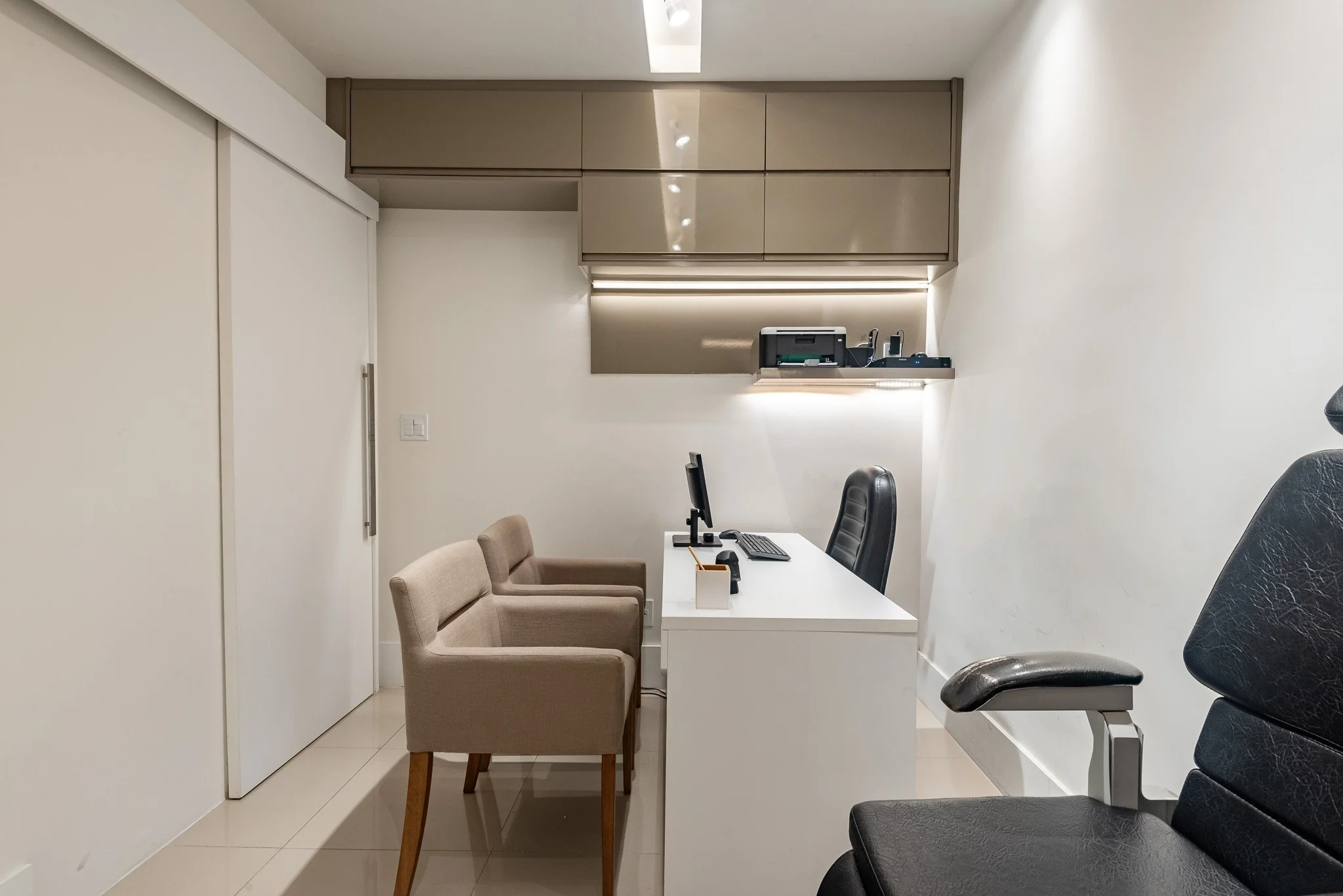 Sala de escritório com mesa branca, duas cadeiras de tecido bege, uma cadeira de escritório preta, um computador e acessórios, armários baixos bege na parede e iluminação embutida.