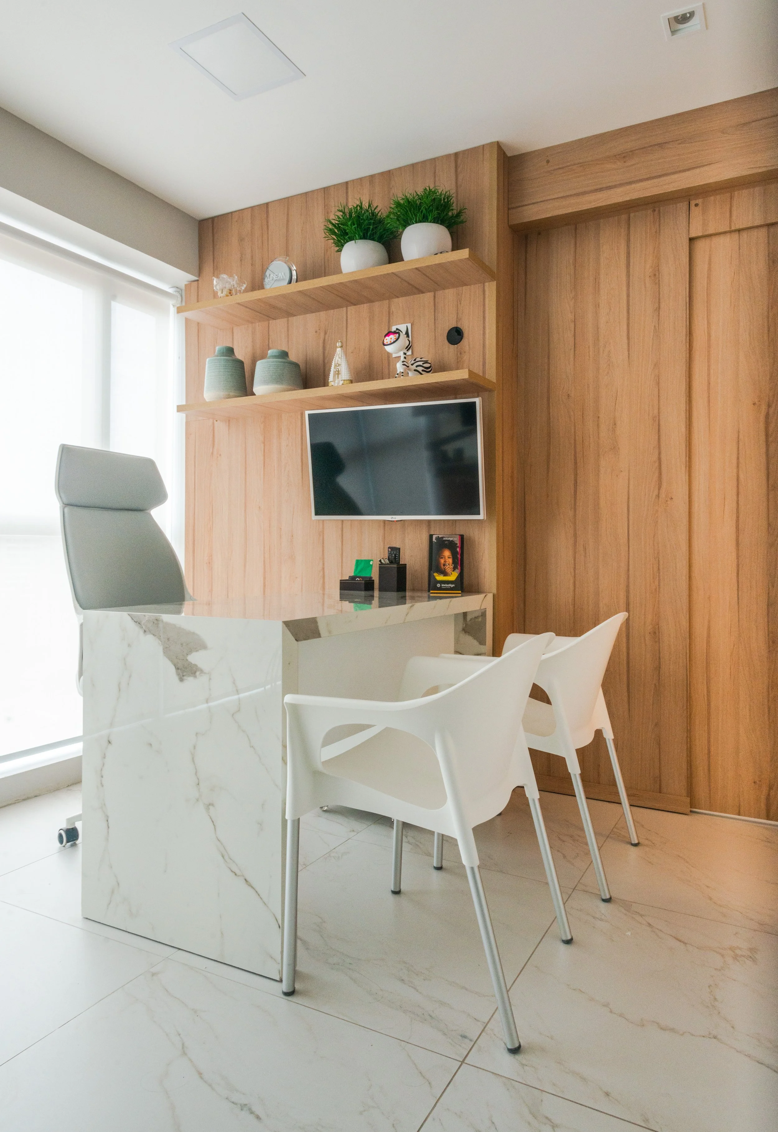 Sala de reunião com mesa de mármore branco, três cadeiras brancas, parede de madeira com prateleiras decorativas, TV montada na parede, e uma grande janela de vidro com cortina branca.