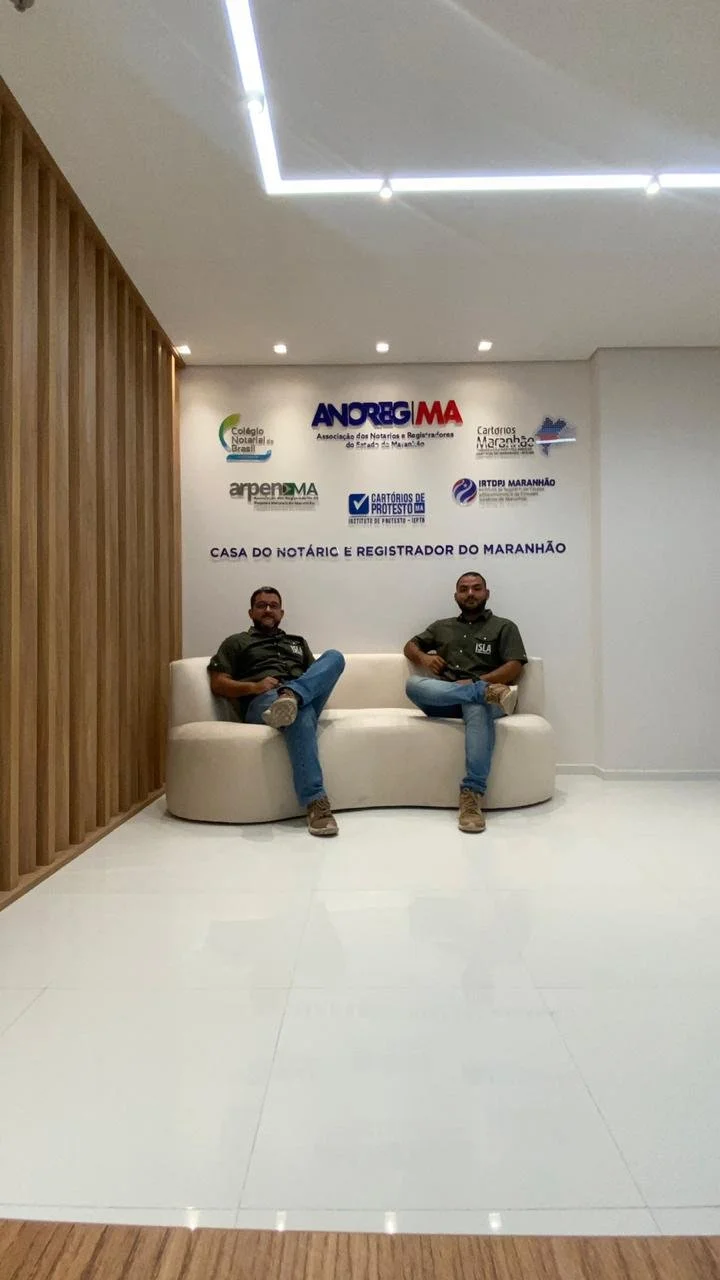 Dois homens sentados em um sofá branco na recepção do Cartório do Maranhão, com parede de fundo mostrando logos de associações e cartórios do Maranhão.