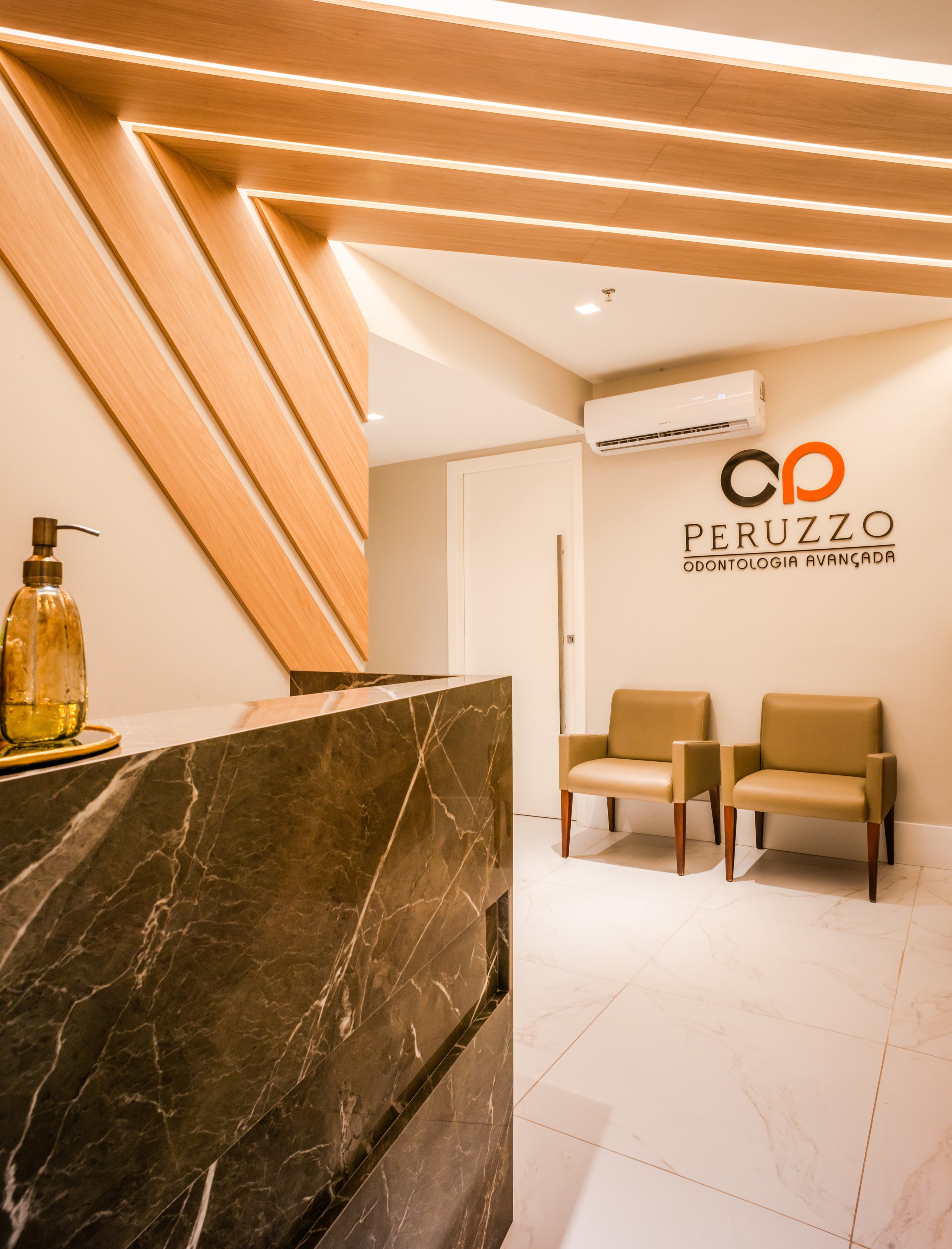 Hall de entrada de uma clínica odontológica com duas cadeiras amareladas no fundo, parede com logo da Peruzzo Odontologia Avançada e decoração moderna com parede de madeira e balcão de mármore escuro.