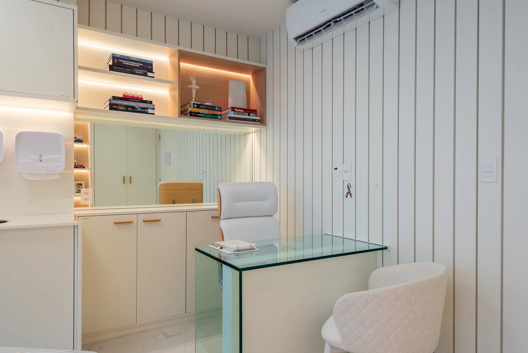 Sala de atendimento com cadeira de escritório branca, mesa de vidro, prateleiras com livros e decoração, espelho grande, paredes com painéis de madeira clara, ar-condicionado no teto, dispensador de papel na parede, ambiente organizado e moderno.