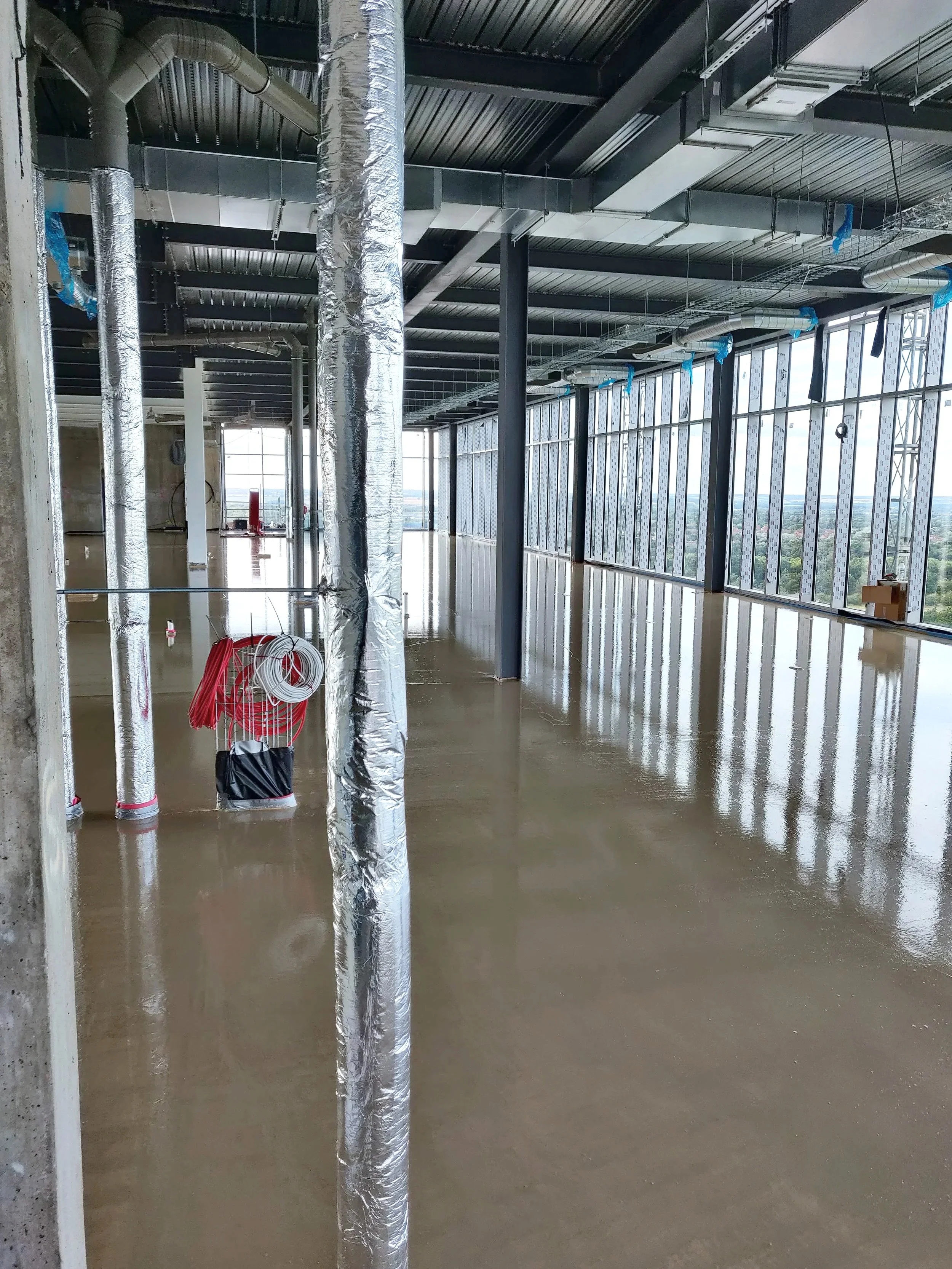 Interior of a high-rise office building under construction with liquid screed floors, exposed ceiling with ductwork, and large windows showing a city view in Buckinghamshire.