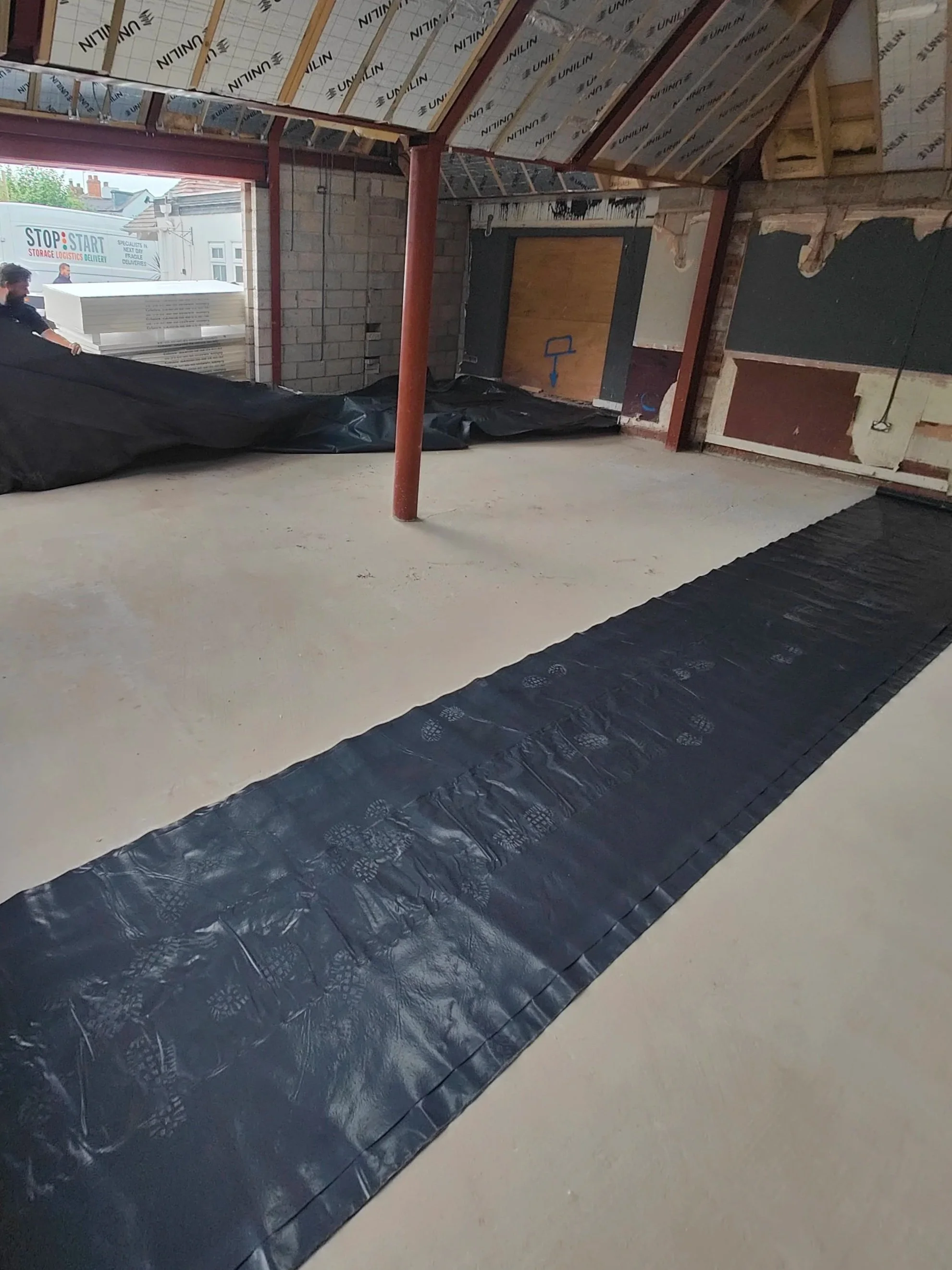 Construction site interior with black damp proof membrane laid on the floor, exposed ceiling with insulation, metal beams, and an open doorway. Worker on the left side handling large black sheet material in Worcestershire.