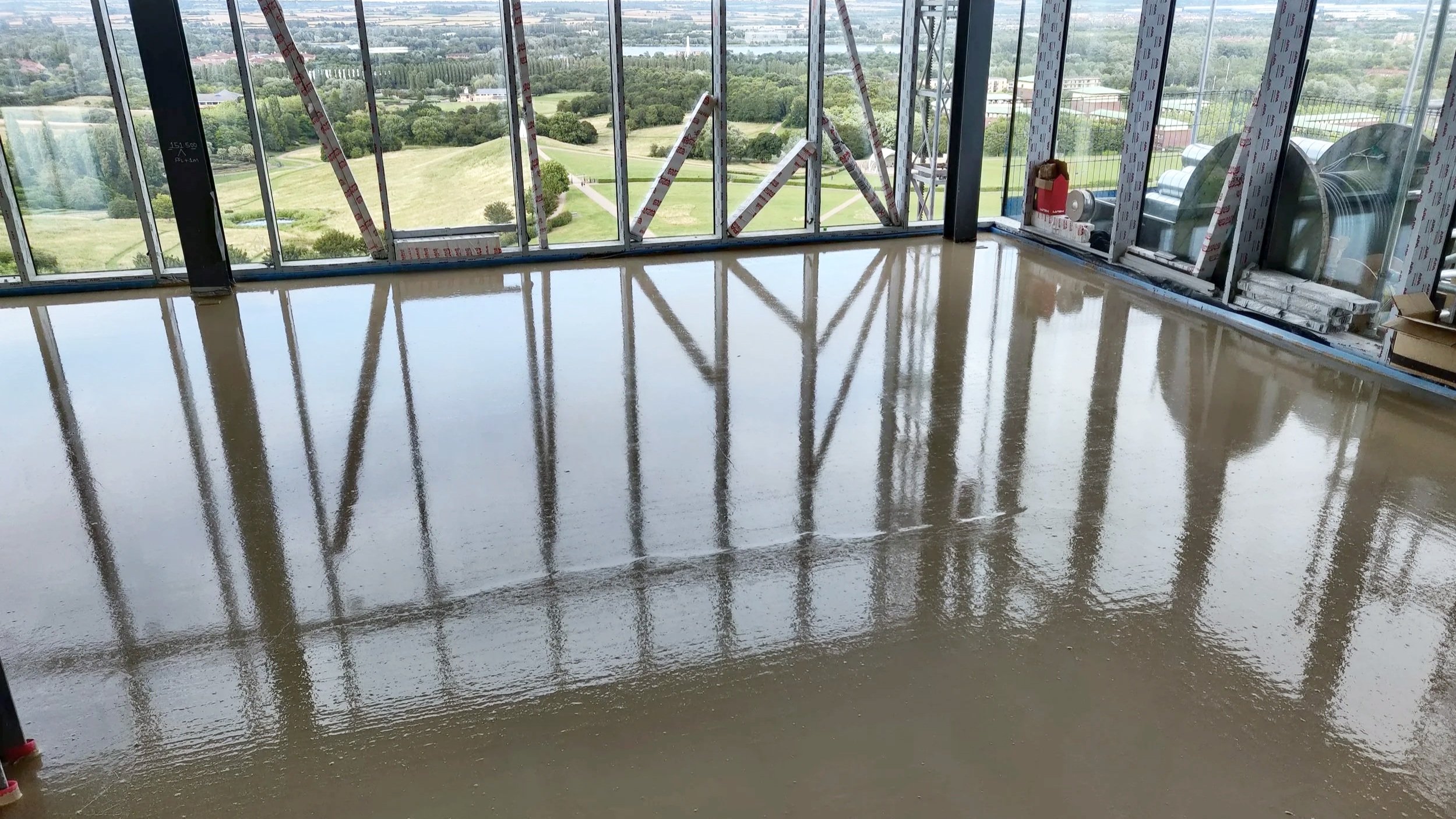 Interior view of a high rise building under construction, with a newly poured liquid screed floor that is shiny and reflective, and large windows showing a green landscape outside in Buckinghamshire.
