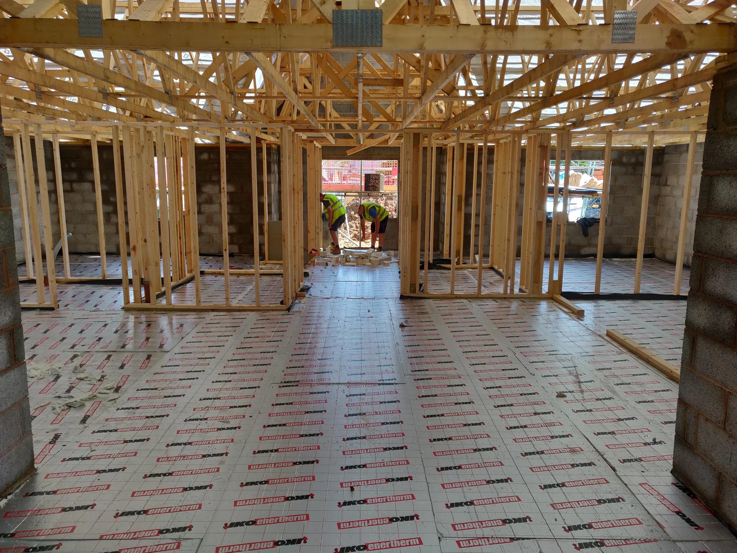 Interior view of a house under construction having insulation installed ready for a liquid screed floor with wooden framing, partially installed roof trusses, and workers in safety vests working near the window opening in Worcestershire.