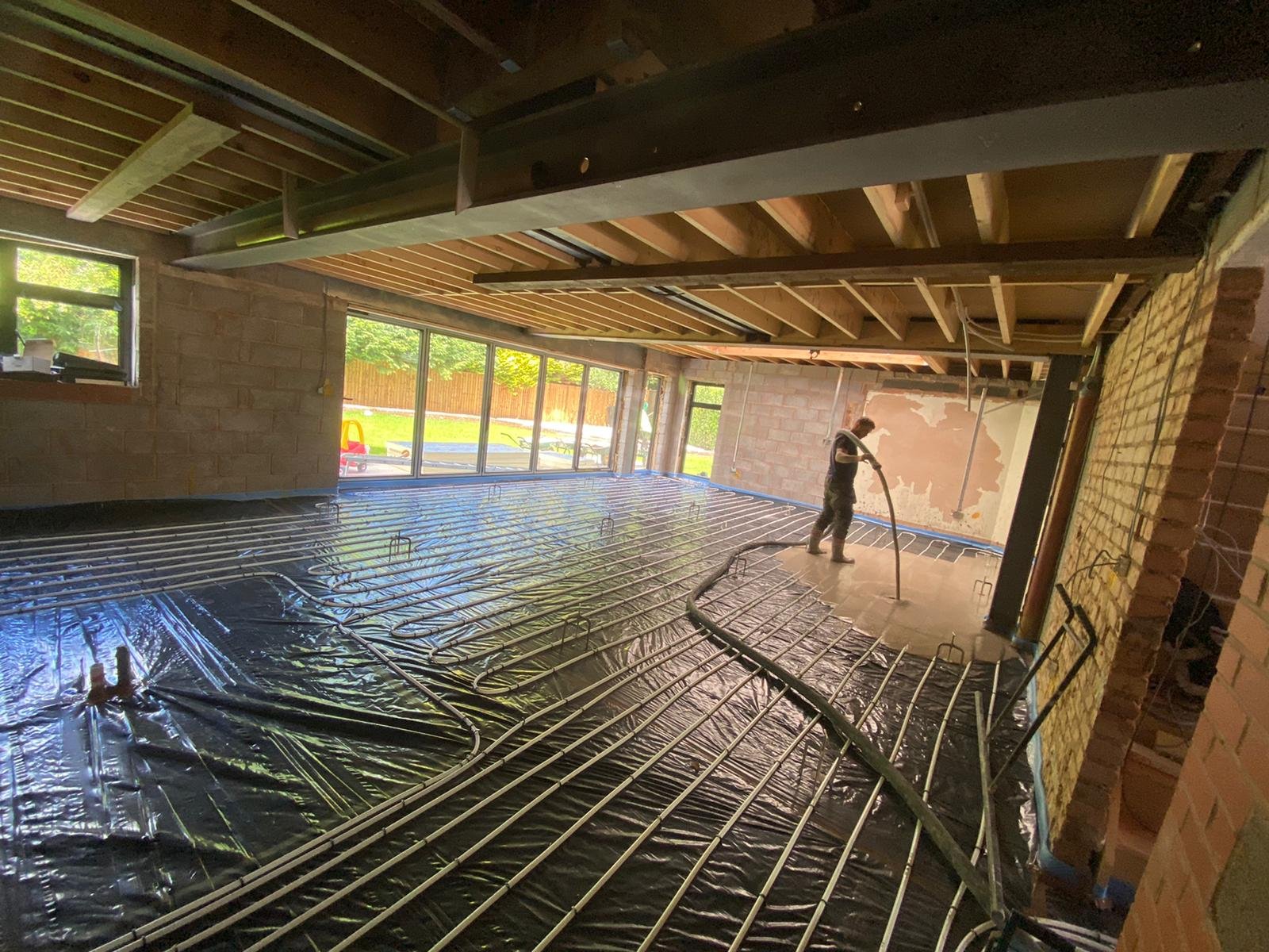 Construction worker installing underfloor heating and liquid screed on the floor of a room under renovation, with large windows revealing a backyard Derbyshire.