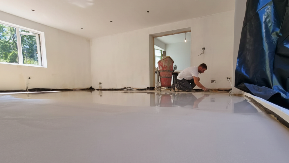 A man installing a self levelling compound floor in a room, with a window showing trees outside in Worcestershire.