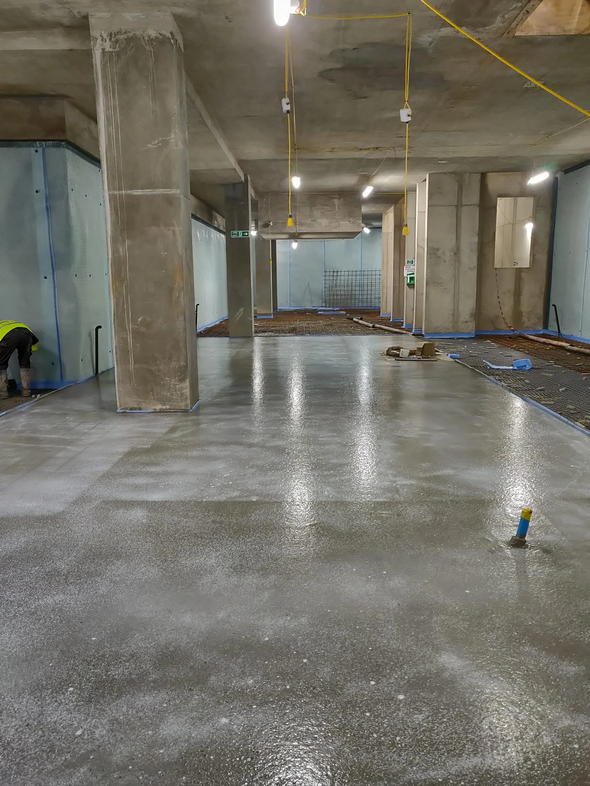 Interior view of a building under construction with a newly poured concrete floor, concrete columns and unfinished walls, with construction worker on the left working on the floor in London