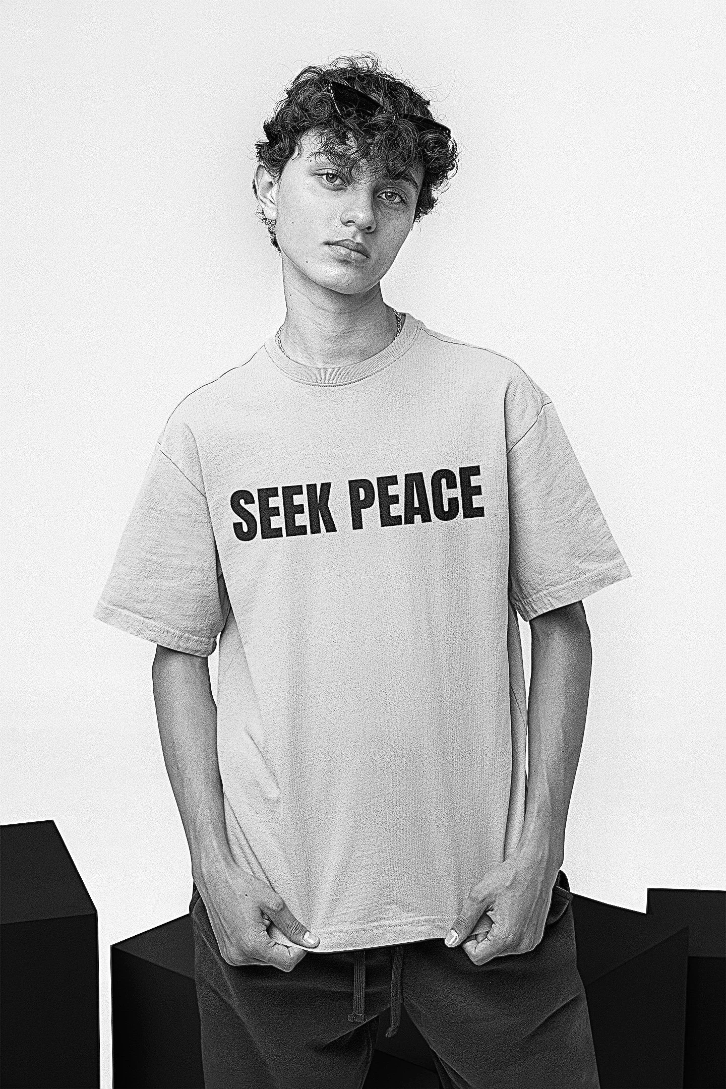 A young person with curly hair wearing a T-shirt that says 'SEEK PEACE' standing against a plain background.