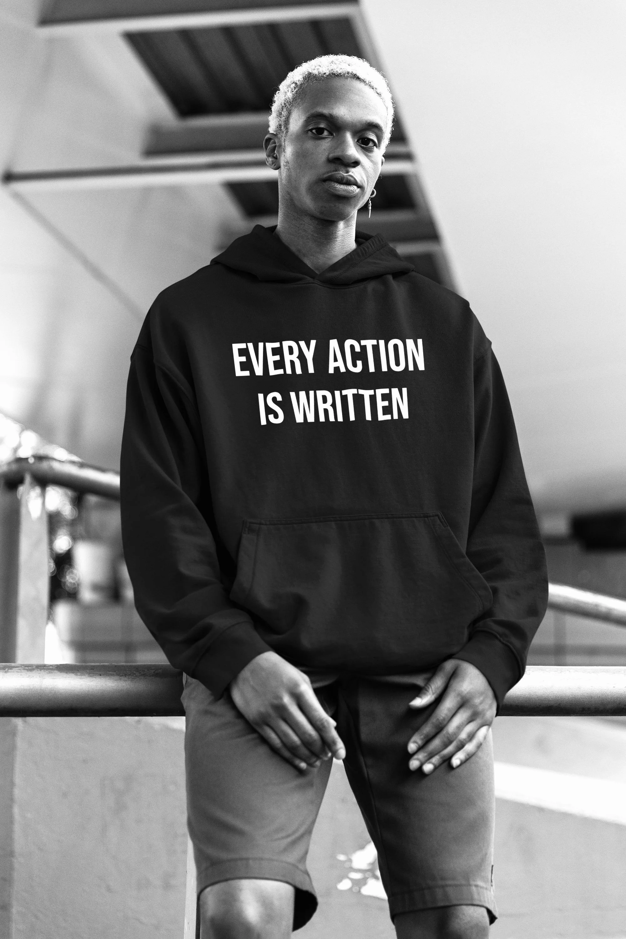 A person with short, light-colored hair wearing a black hoodie with the text 'EVERY ACTION IS WRITTEN' printed on front, leaning against a railing outdoors.