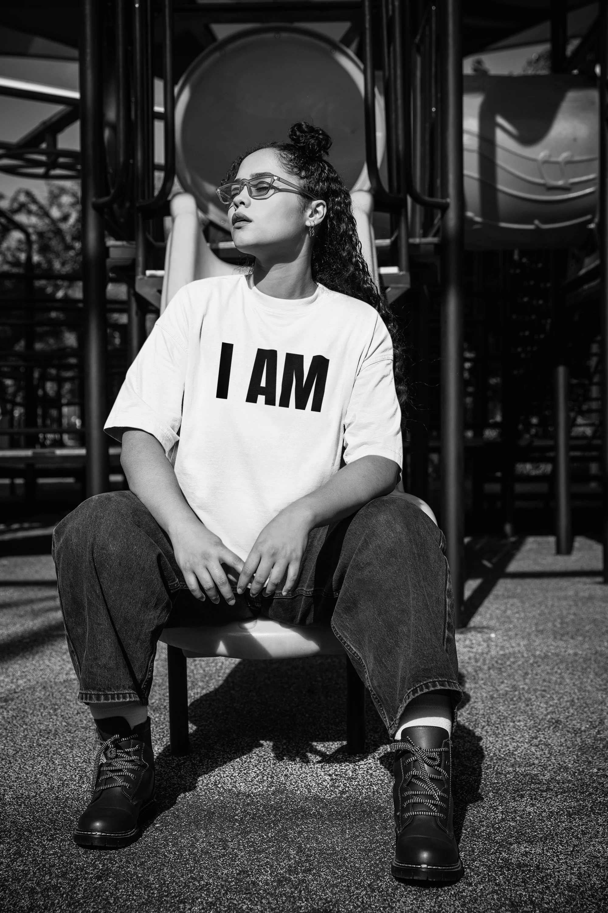 A woman sitting on a small chair at a playground, wearing glasses, a white T-shirt with "I AM" printed on it, baggy jeans, and boots, with her eyes closed and head tilted slightly upward, in black and white.