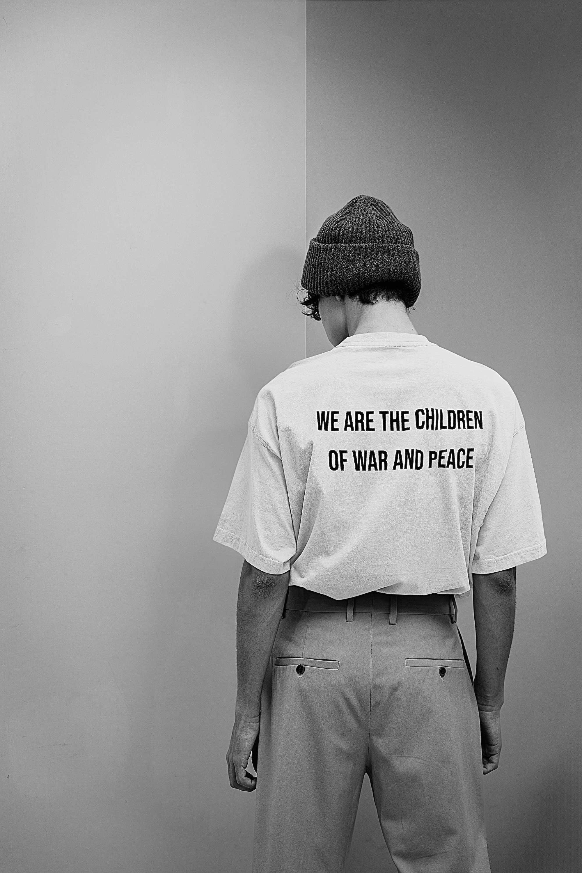 Person wearing a beanie and a T-shirt with the message "We are the children of war and peace" on the back, standing against a plain wall.