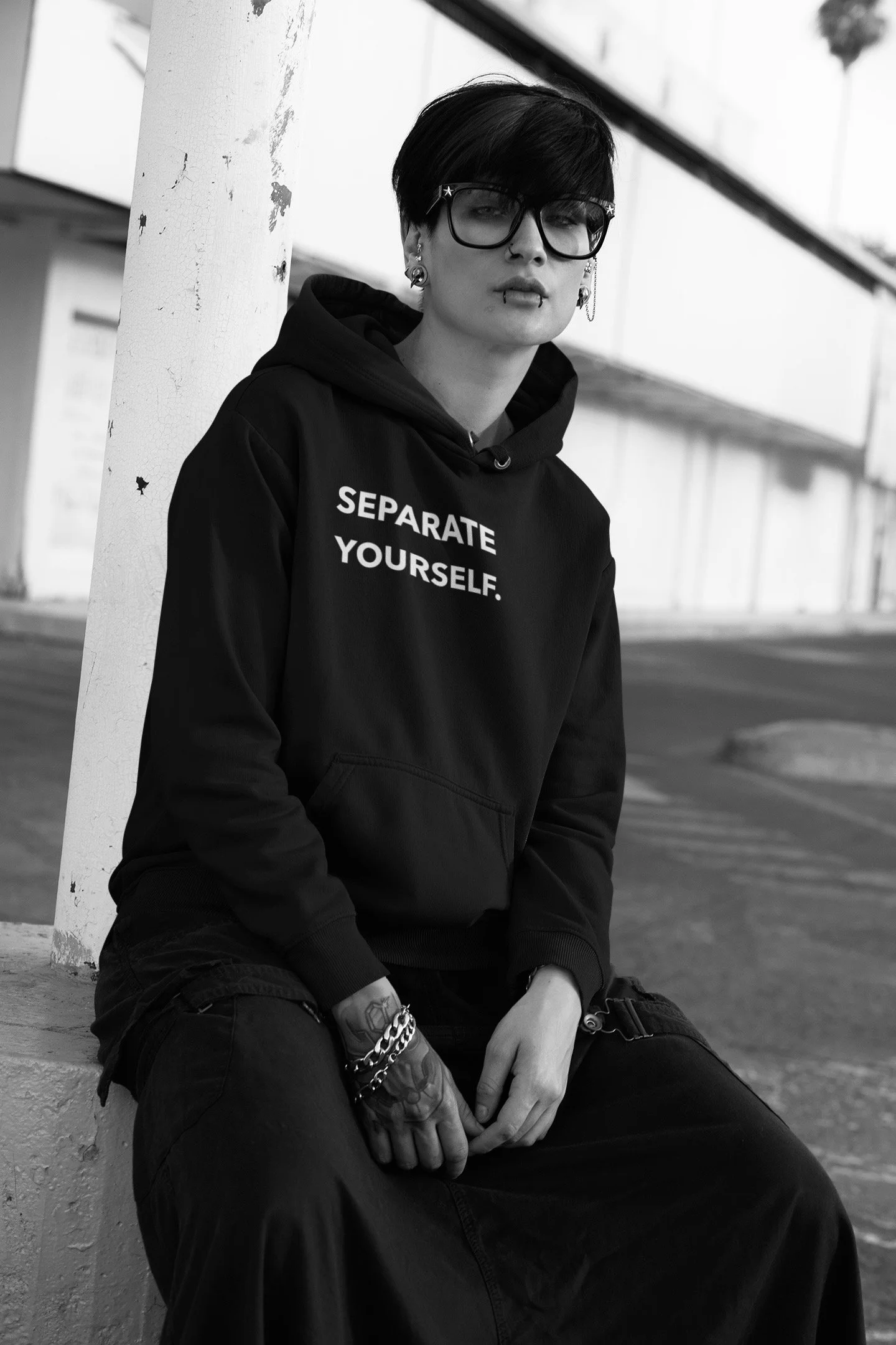 A person with short dark hair and glasses sitting on a curb, wearing a hoodie that says "SEPARATE YOURSELF." in white text, and various jewelry, in an urban setting.