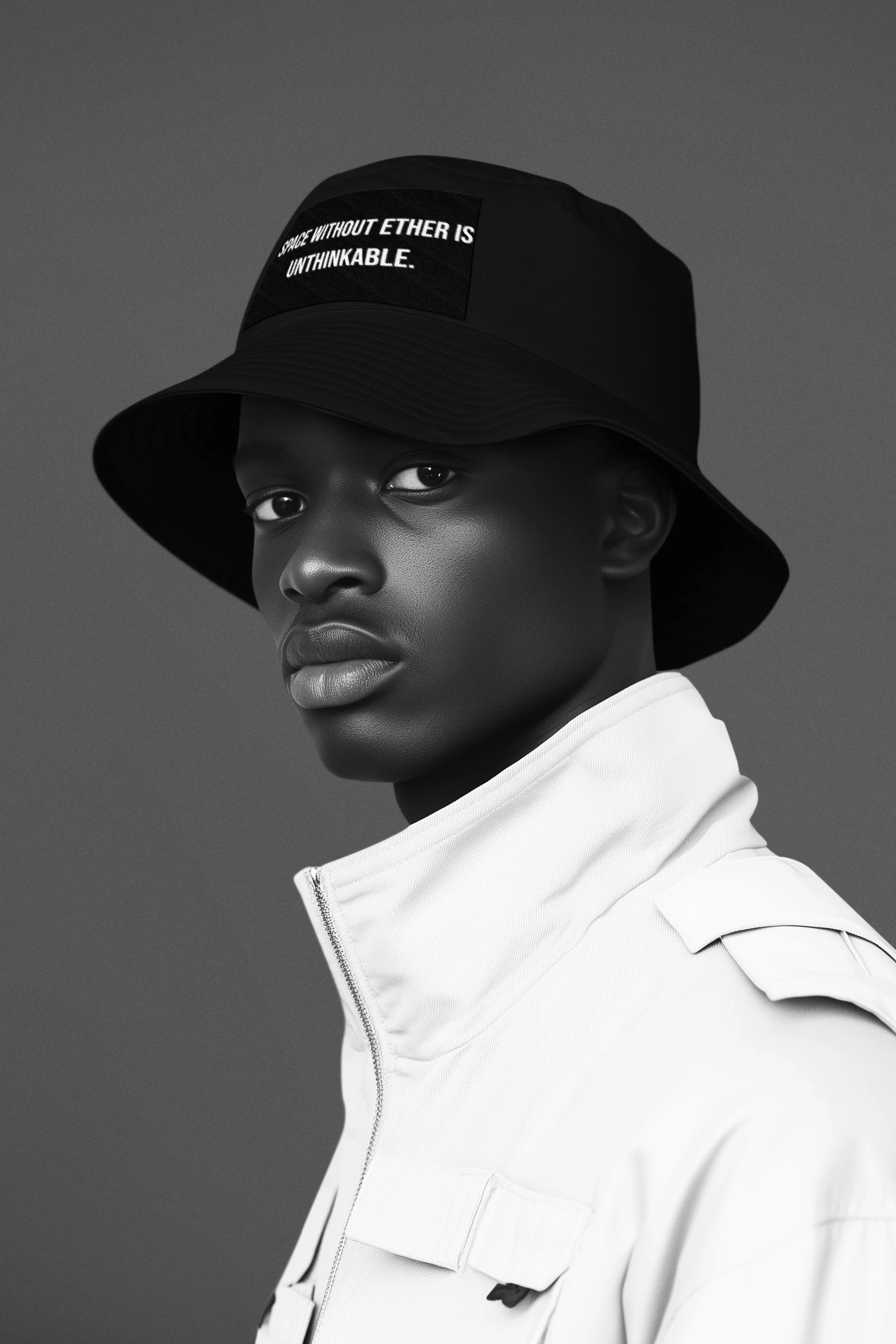 A young Black man wearing a black bucket hat with a quote and a white jacket, looking at the camera in a studio with a plain background.