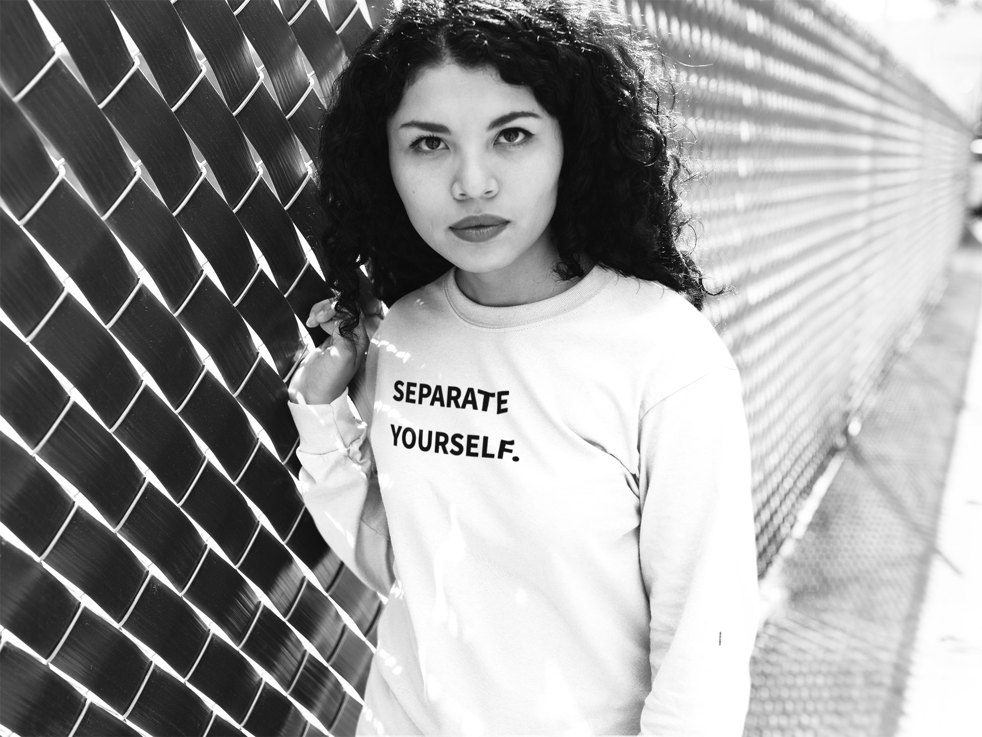 A young woman with curly hair standing next to a chain-link fence, holding onto the fence with one hand, wearing a shirt that says 'SEPARATE YOURSELF.' in bold letters.