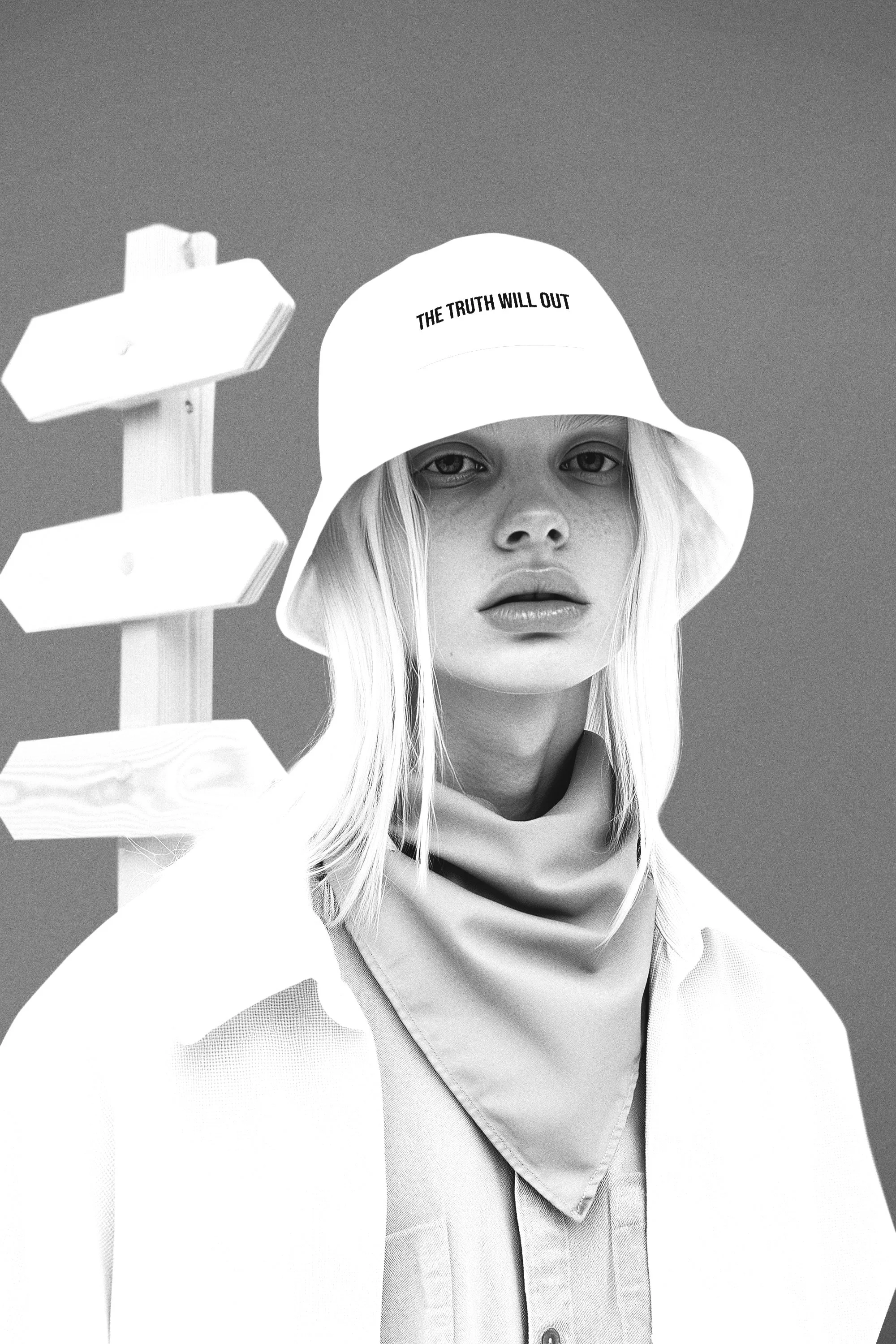Black and white photo of a woman with long light-colored hair wearing a bucket hat with the text "THE TRUTH WILL OUT" and a bandana around her neck, standing next to a wooden signpost with blank directional signs.