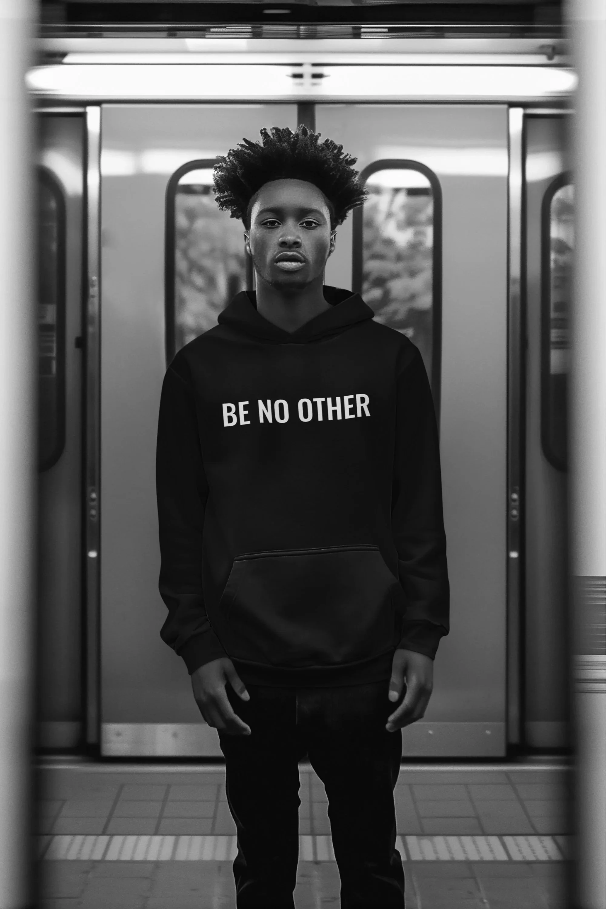 A young man standing on a train platform wearing a black hoodie with the words 'BE NO OTHER' printed on the front, framed by the open doors of a subway train.