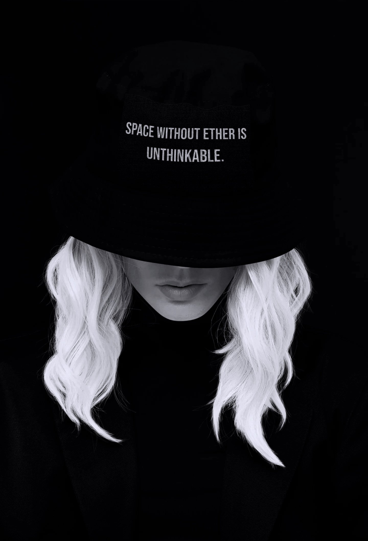 A woman with platinum blonde hair wearing a black hat with the words 'Space Without Ether Is Unthinkable' embroidered on it, and a dark outfit, set against a black background.