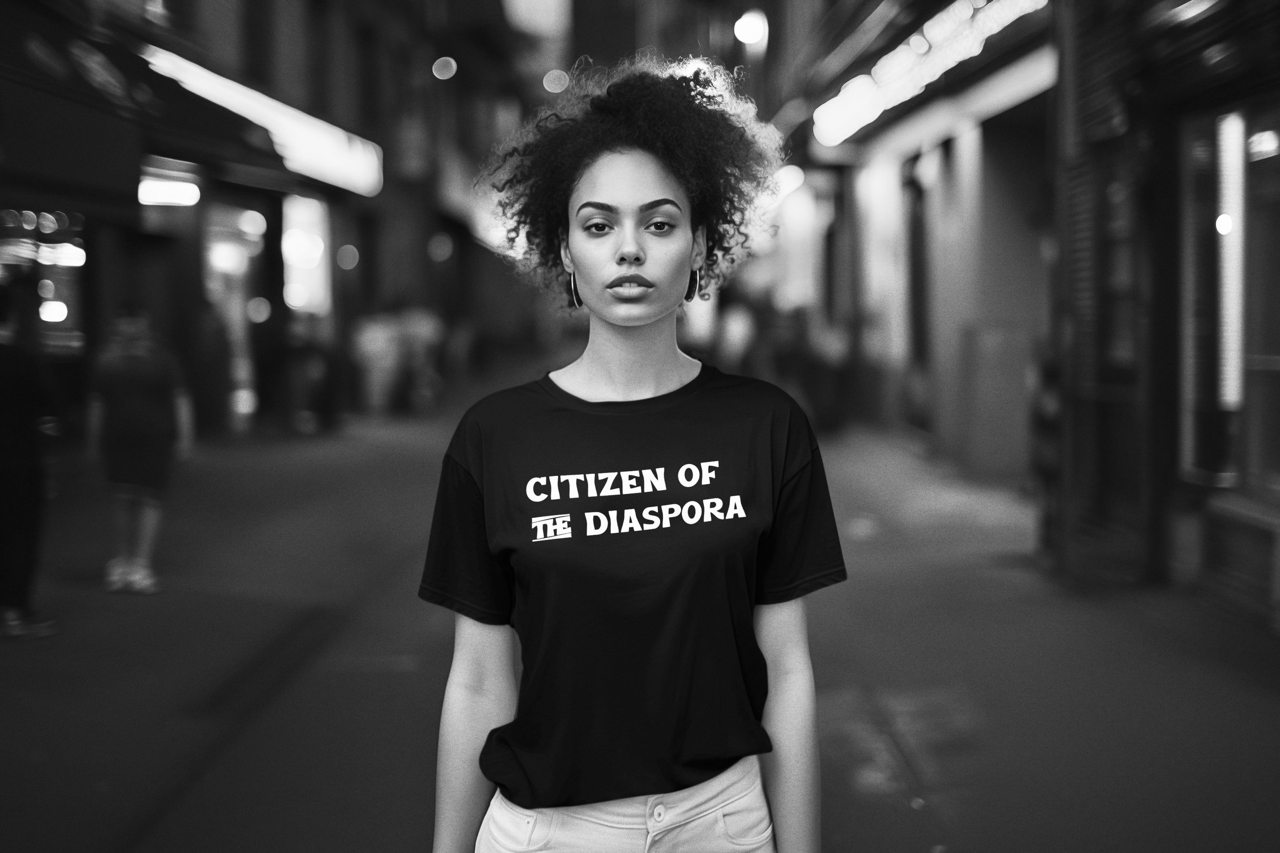 A young woman with curly hair wearing a black T-shirt that says "Citizen of the Diaspora" in bold letters, standing on a city street at night.
