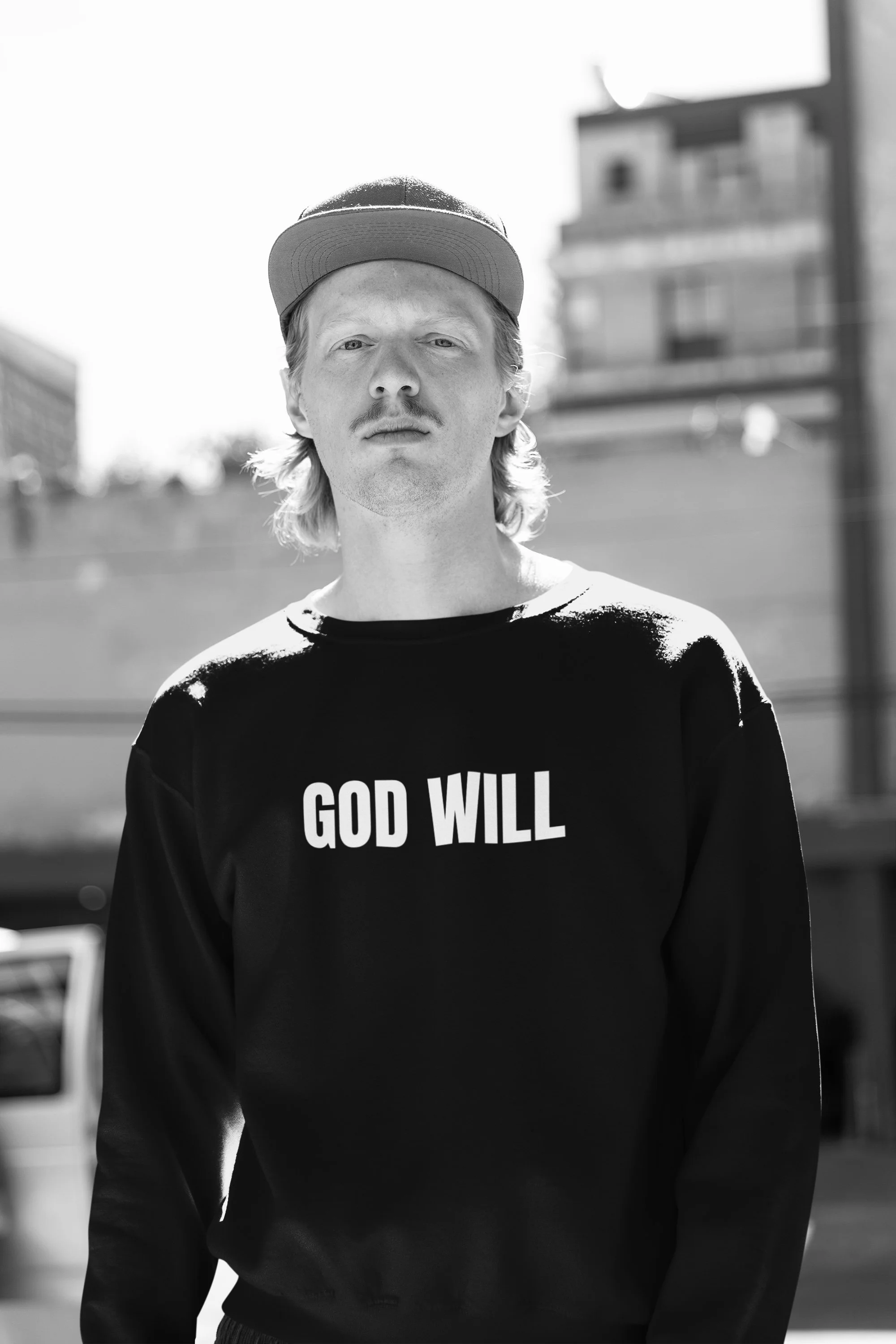 Black and white photo of a young man wearing a cap and sweatshirt that reads "GOD WILL" standing outdoors in an urban setting.
