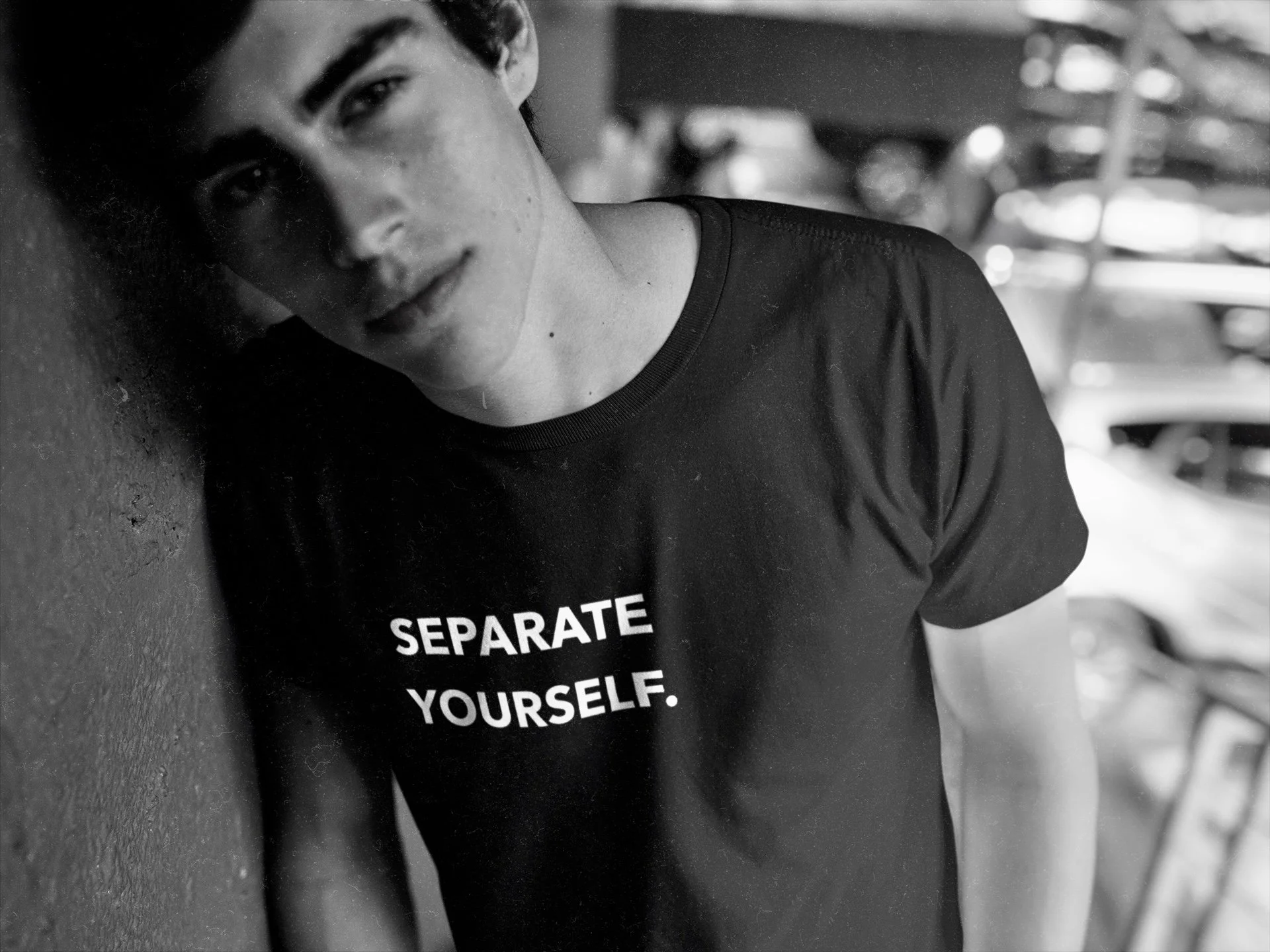 A young man with dark hair leaning against a textured wall in a parking garage, wearing a black T-shirt with white text that reads "SEPARATE YOURSELF." The background shows blurred parked cars.