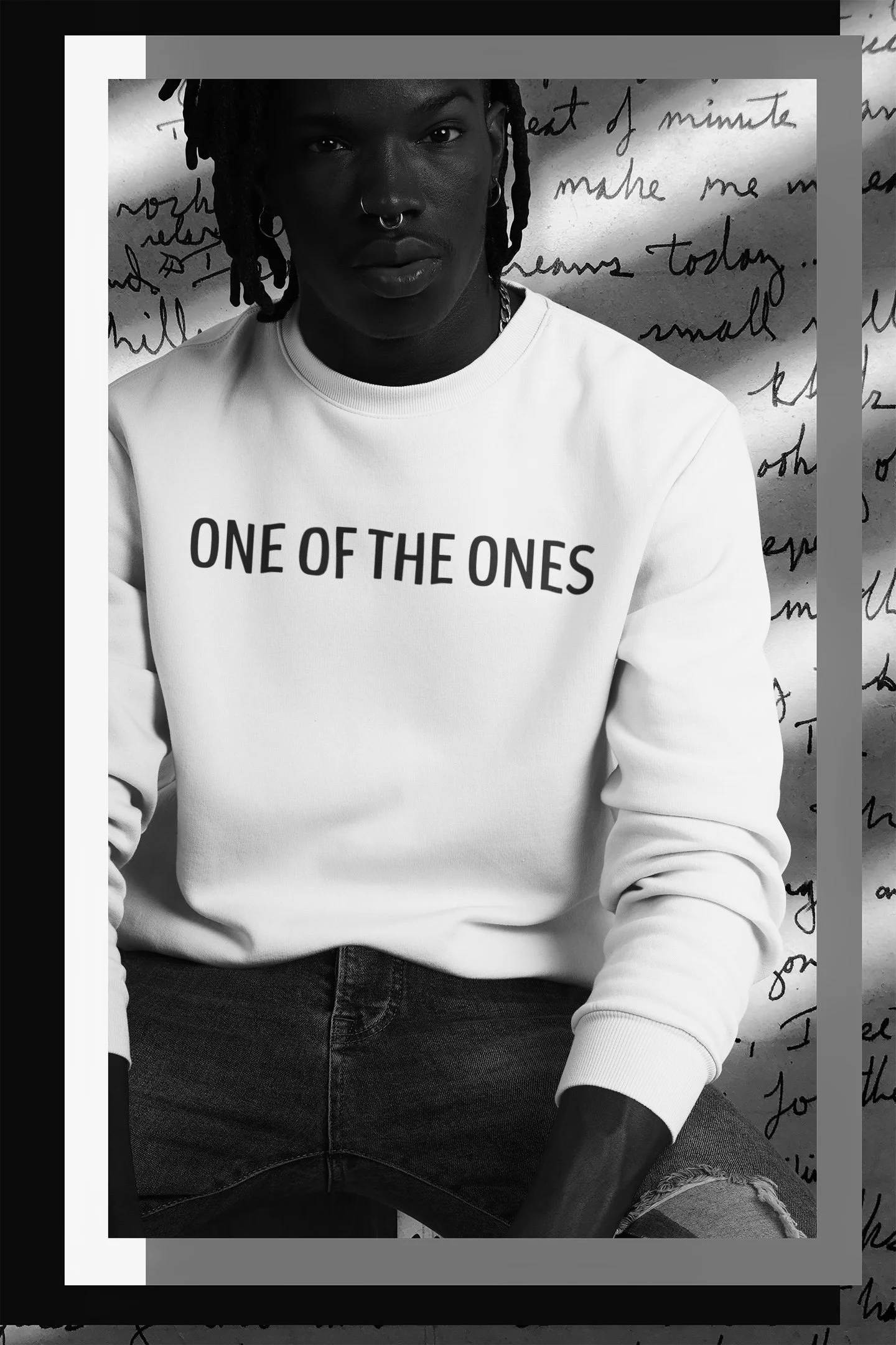 A person with dark skin and dreadlocks wearing a white hoodie with the words "ONE OF THE ONES" printed on it, sitting against a handwritten letter backdrop.