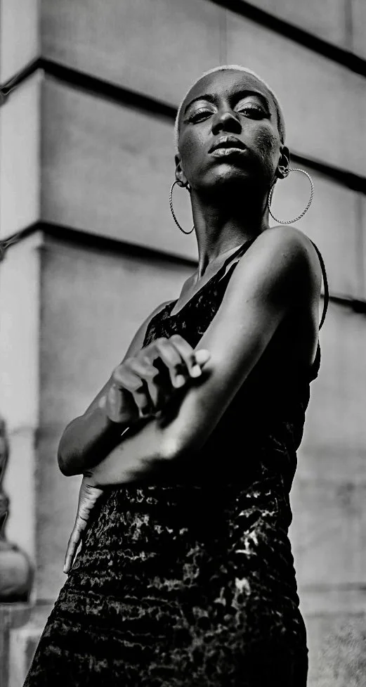 A black and white photo of a confident woman with short hair, wearing large hoop earrings and a leopard-print dress, standing outdoors against a modern architectural background.