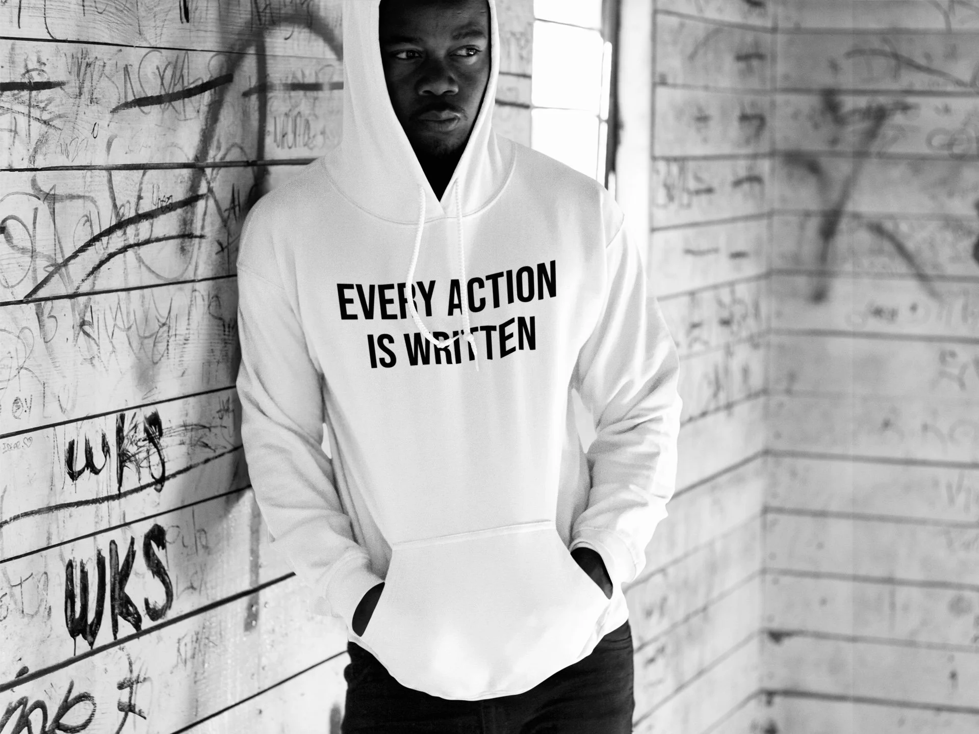 A young man in a white hoodie with the text "Every Action is Written" stands with hands in pockets against a graffiti-covered wall in black and white.