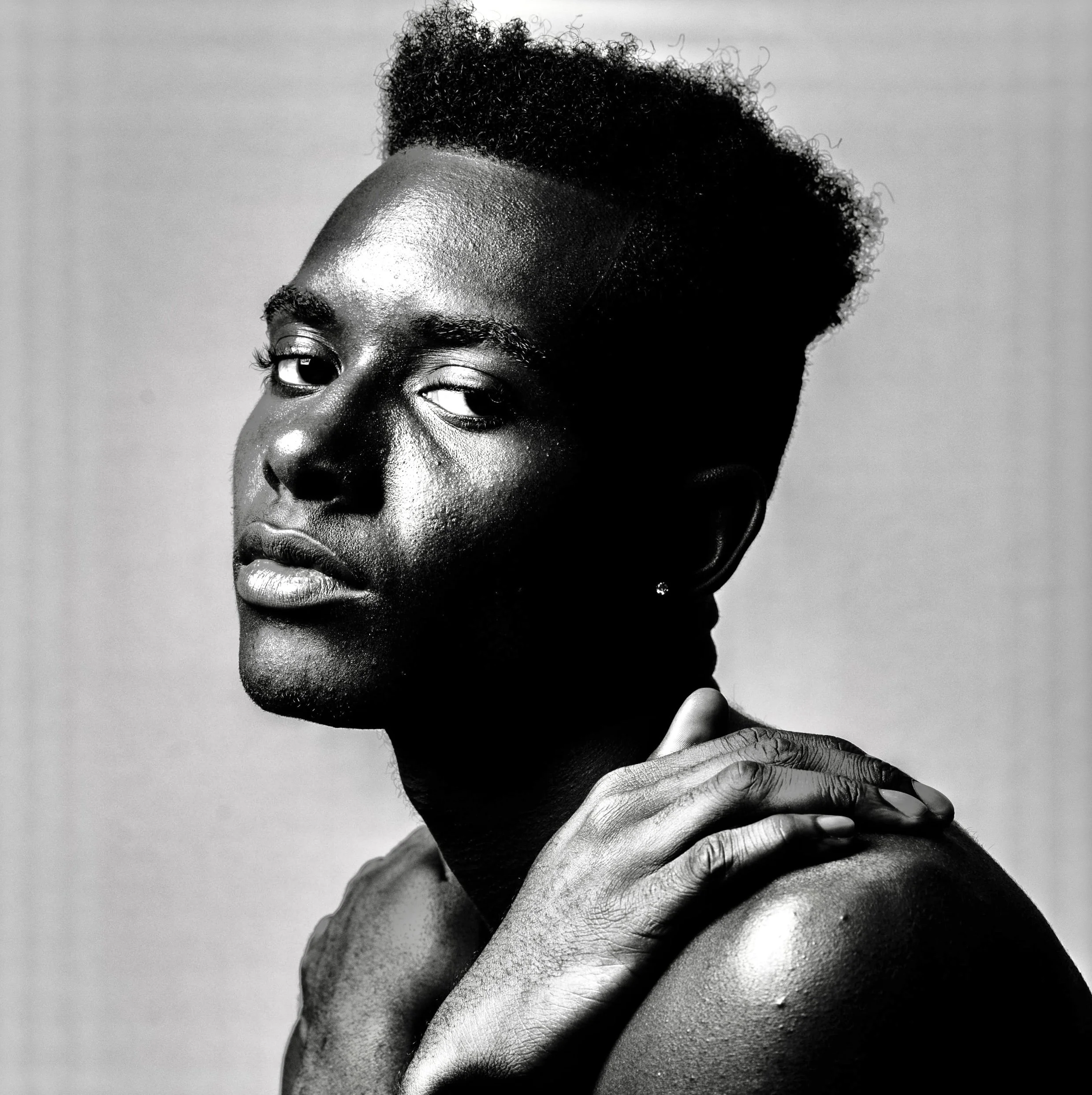Black and white portrait of a person with short, curly hair, touching their shoulder, with intense facial expression.