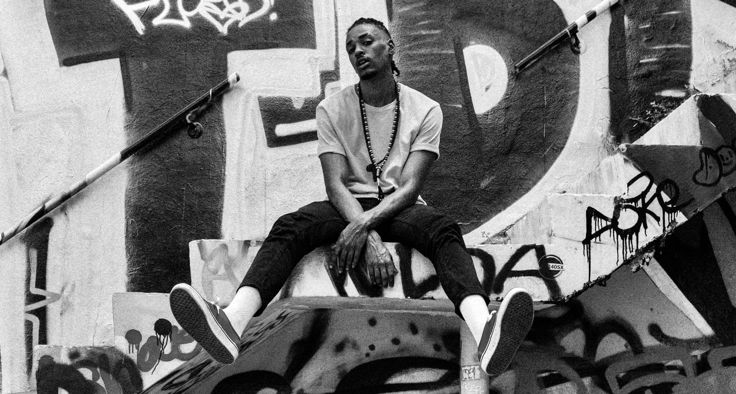 A young man sitting on graffiti-covered stairs in an urban setting, wearing a t-shirt, dark pants, sneakers, and a beaded necklace, with one arm resting on his knee and the other hand on his foot, looking off to the side.