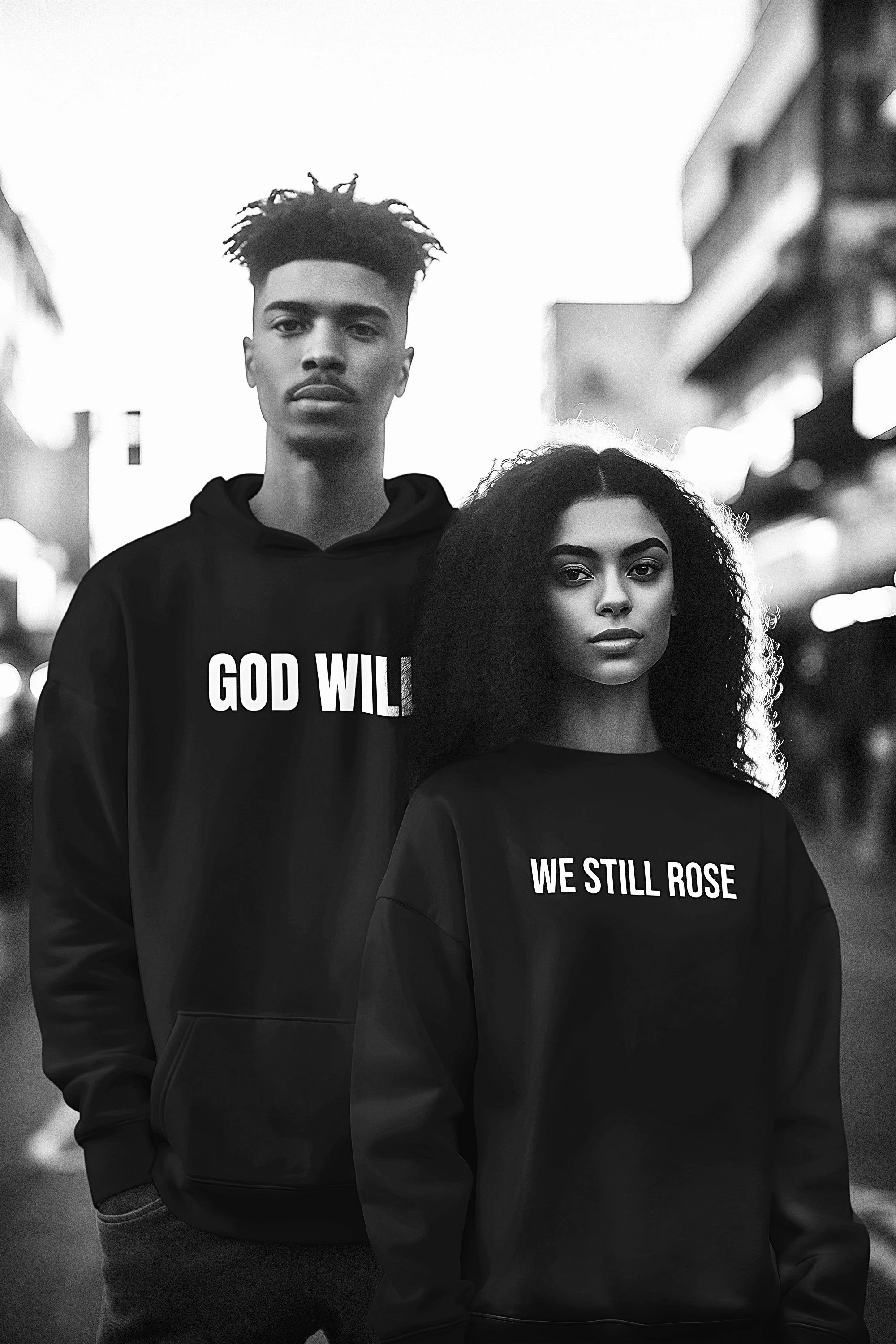 A black and white photo of a young man and woman standing outdoors in an urban environment at dusk. The man has short tropical curly hair and wears a hoodie that says 'GOD WILL' while the woman has voluminous curly hair and wears a sweatshirt that sa